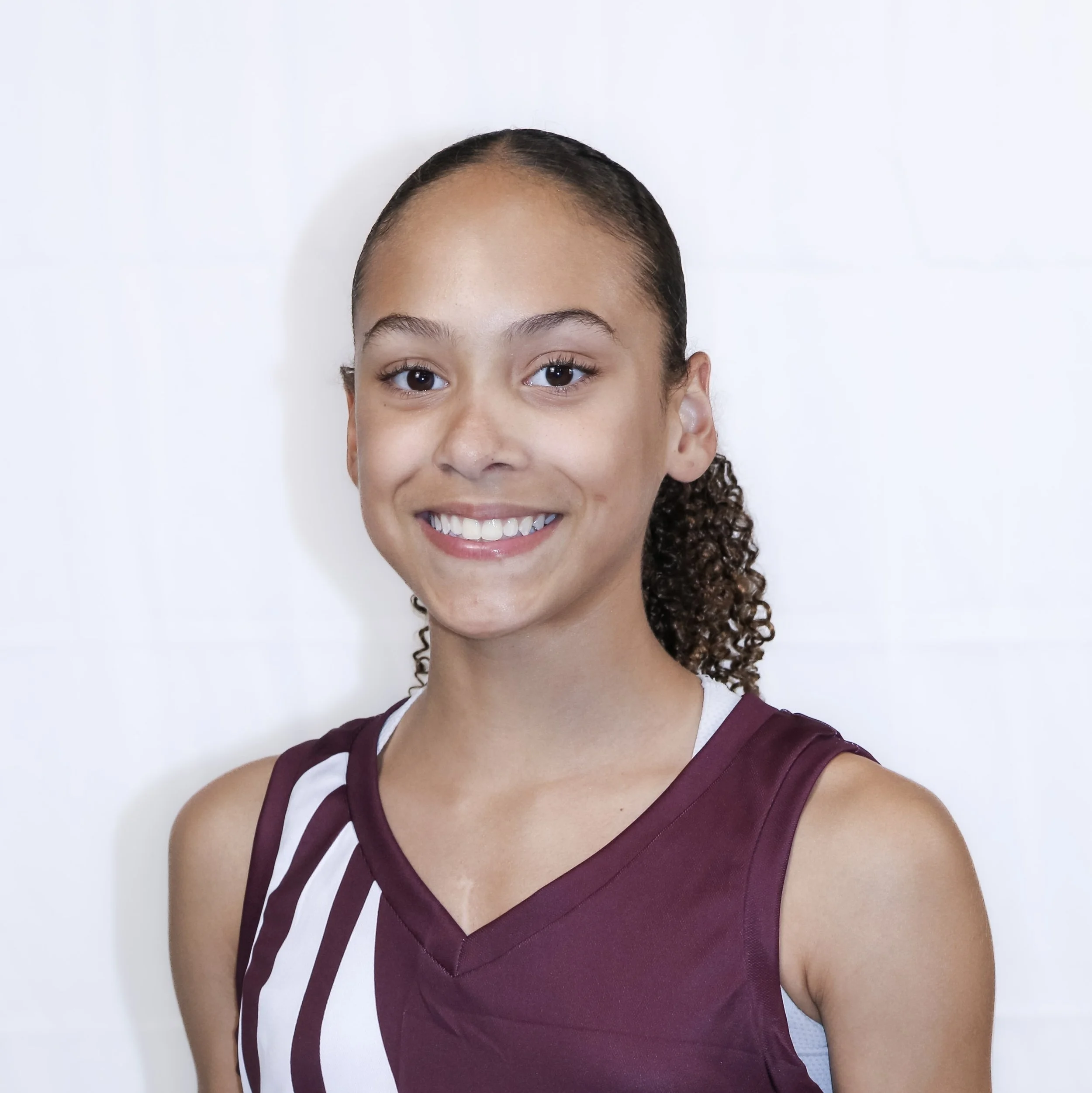 Close-up of a young woman with curly hair and a bright smile wearing a maroon and white sports uniform against a plain white background.