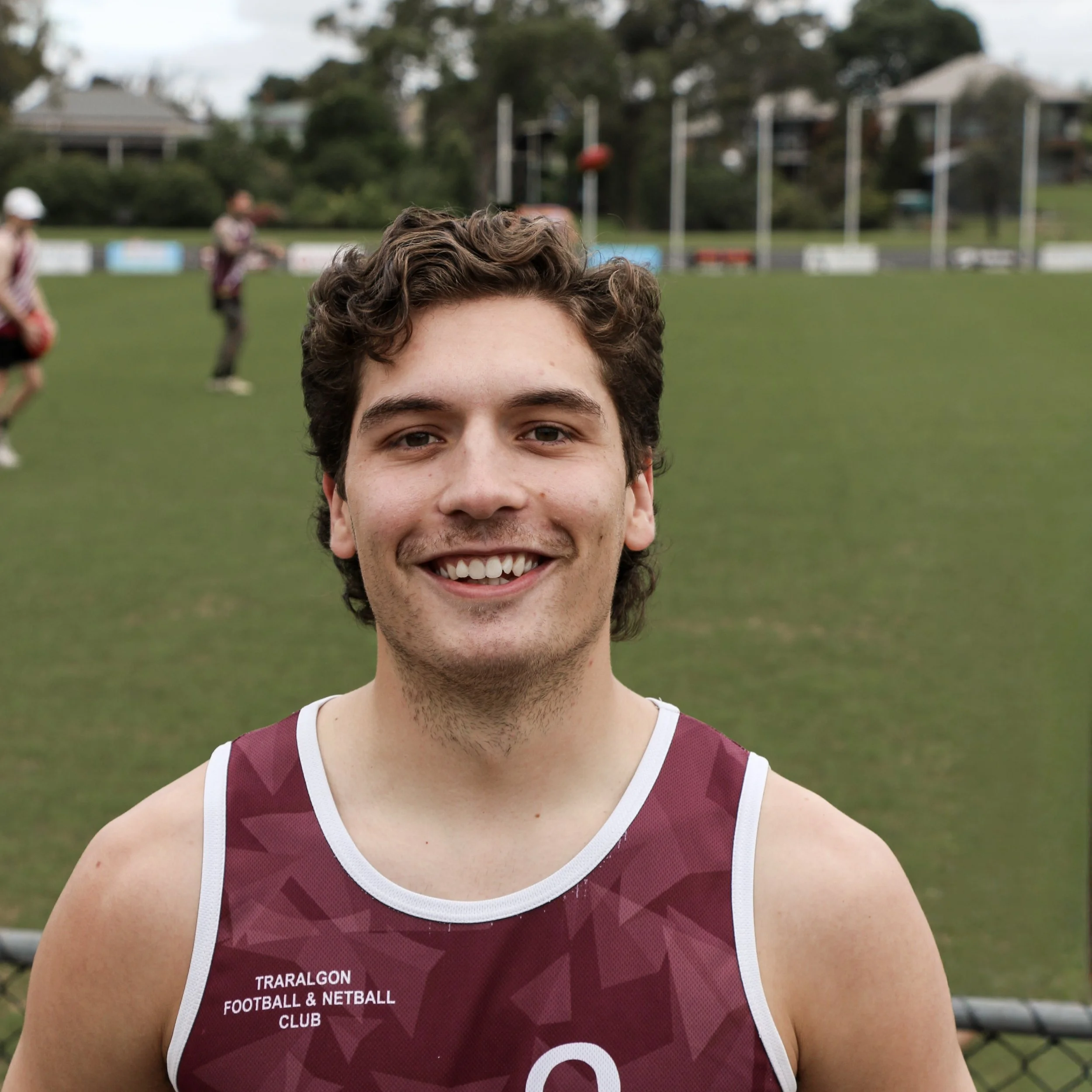 Young man in sports jersey smiling on a field, with other players practicing in the background.