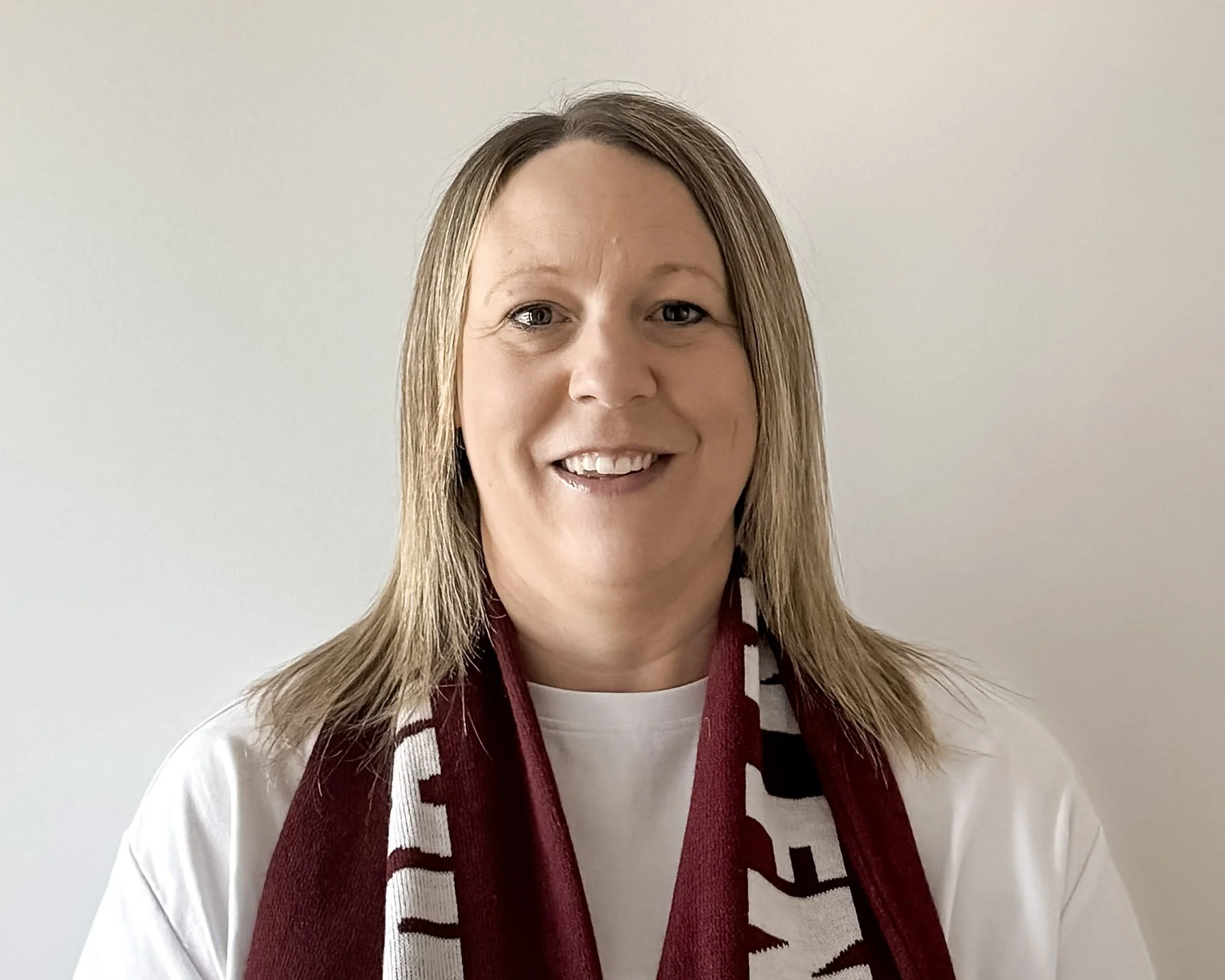 A woman with light brown hair smiling, wearing a maroon and white scarf over a white shirt, standing against a plain white wall.