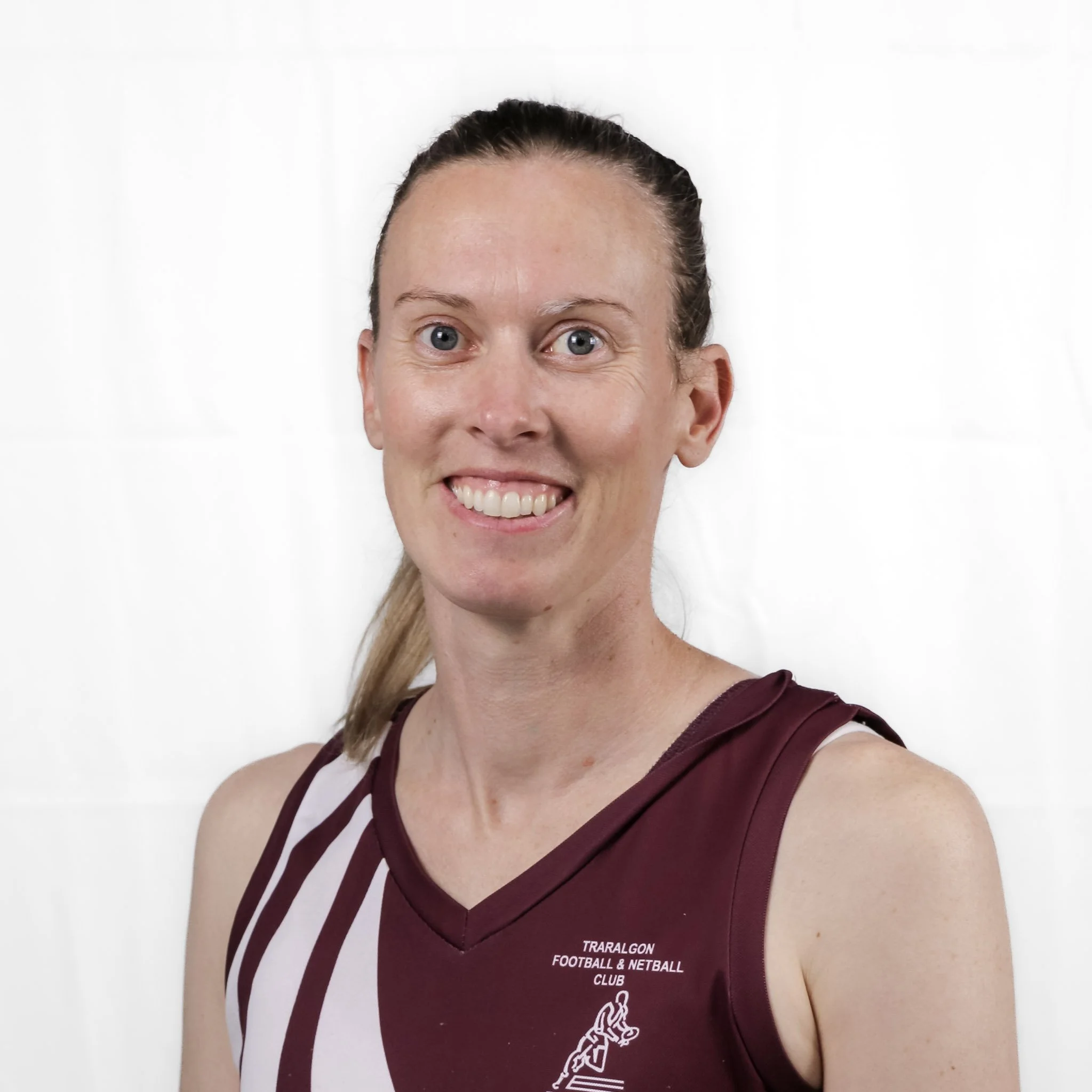 Close-up of a woman with light skin, blue eyes, and brown hair tied back, smiling and wearing a maroon sports uniform with "TRARALAGON FOOTBALL & NETBALL CLUB" text.