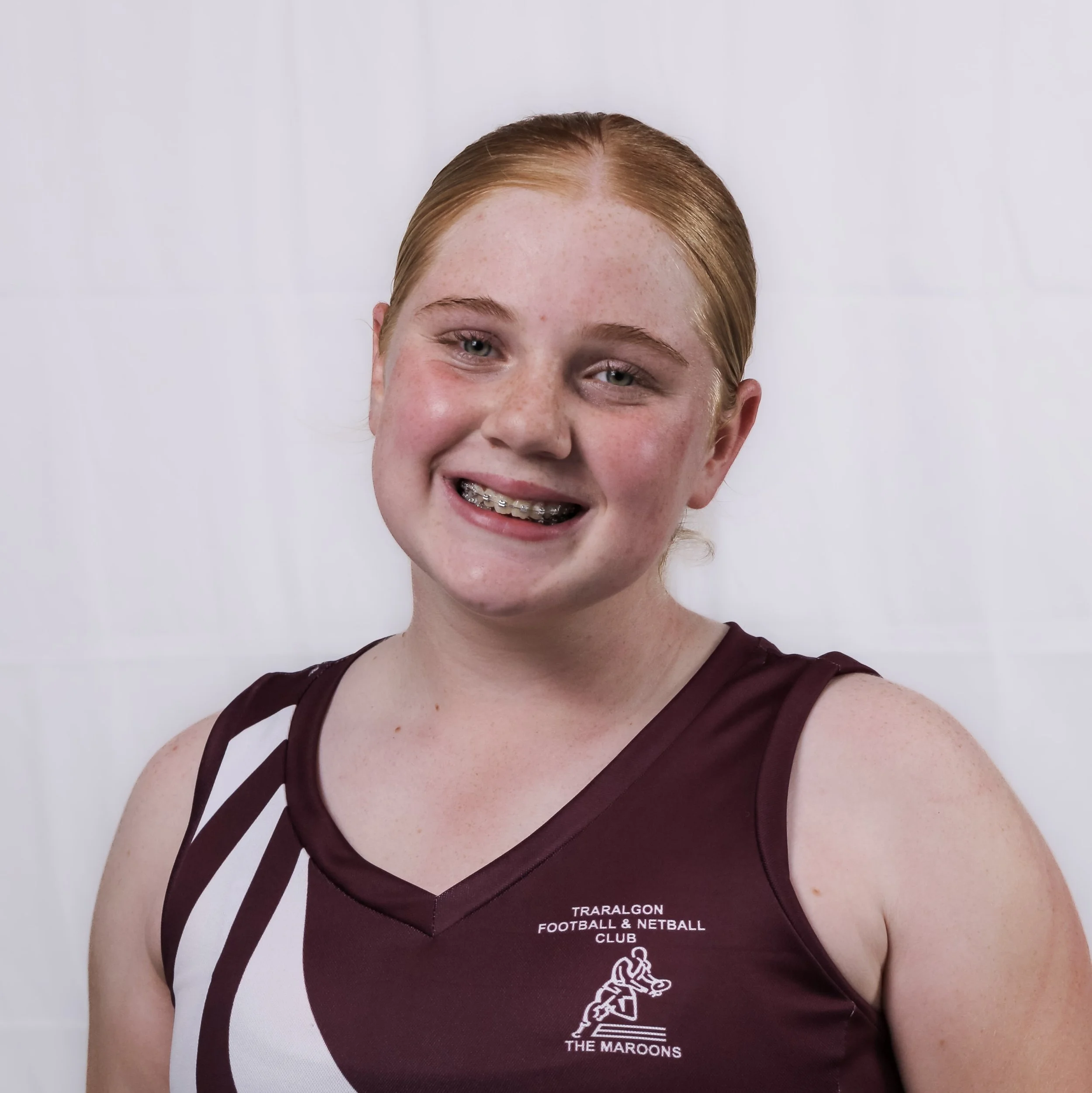 A person with red hair and freckles smiling and wearing a maroon sports jersey with white stripes, representing the Traralgon Football and Netball Club.