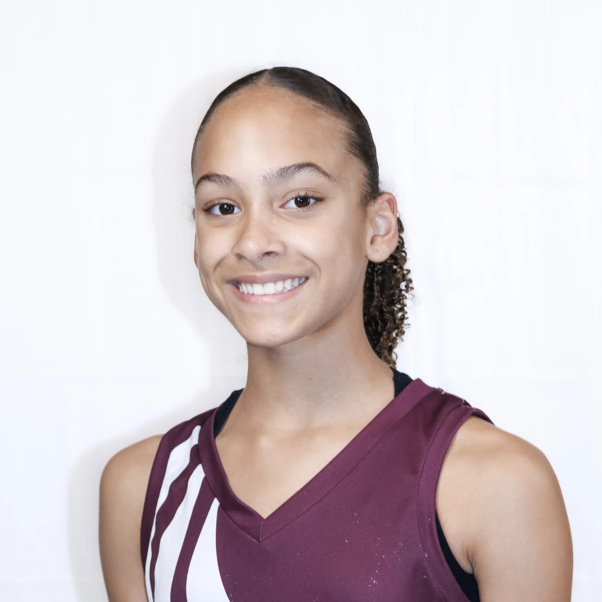 Close-up of a young woman with light brown skin, curly hair tied back, smiling, wearing a maroon basketball jersey with white accents, standing against a plain white background.