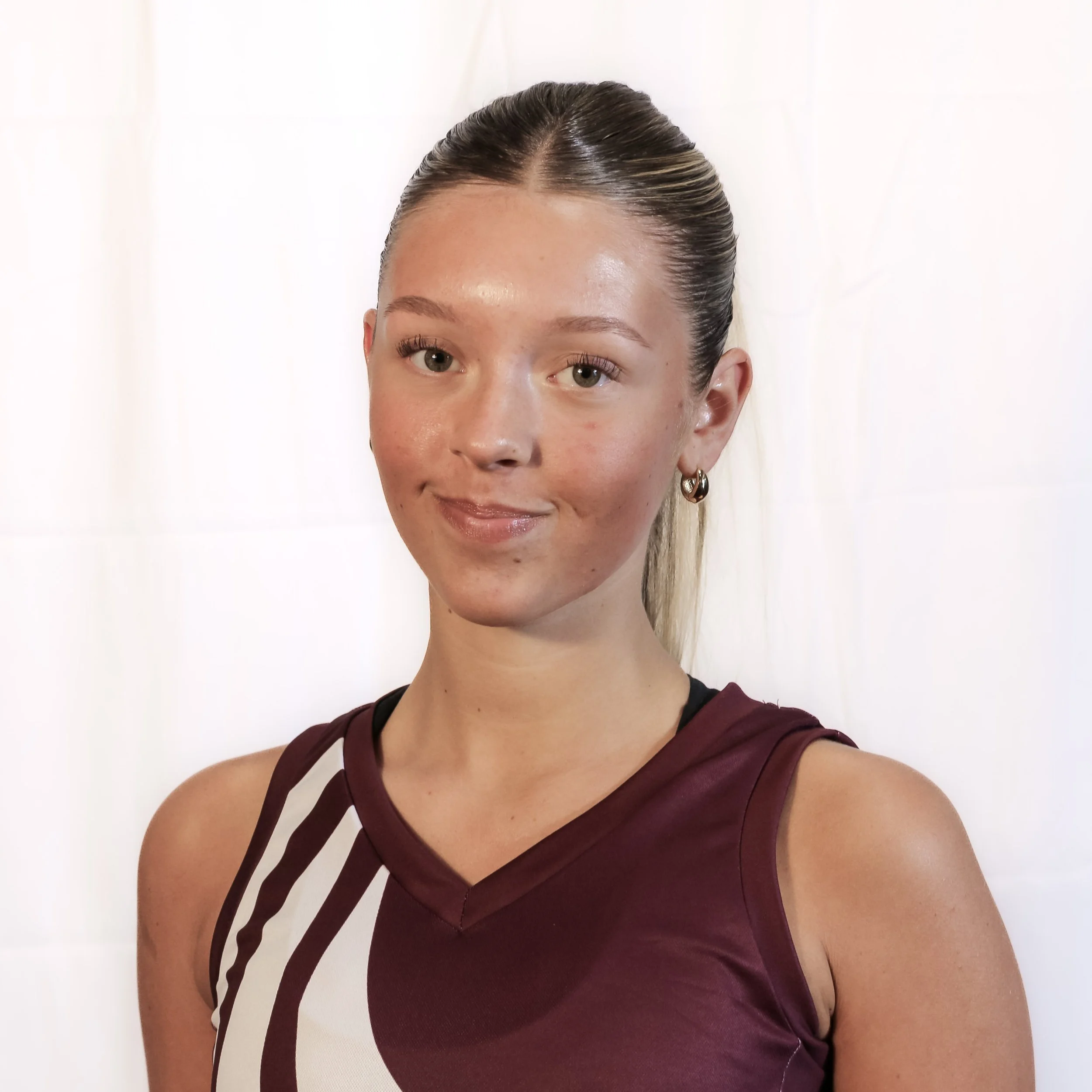 Close-up of a woman with her hair pulled back, wearing a maroon sleeveless top with black straps, standing against a plain white background.