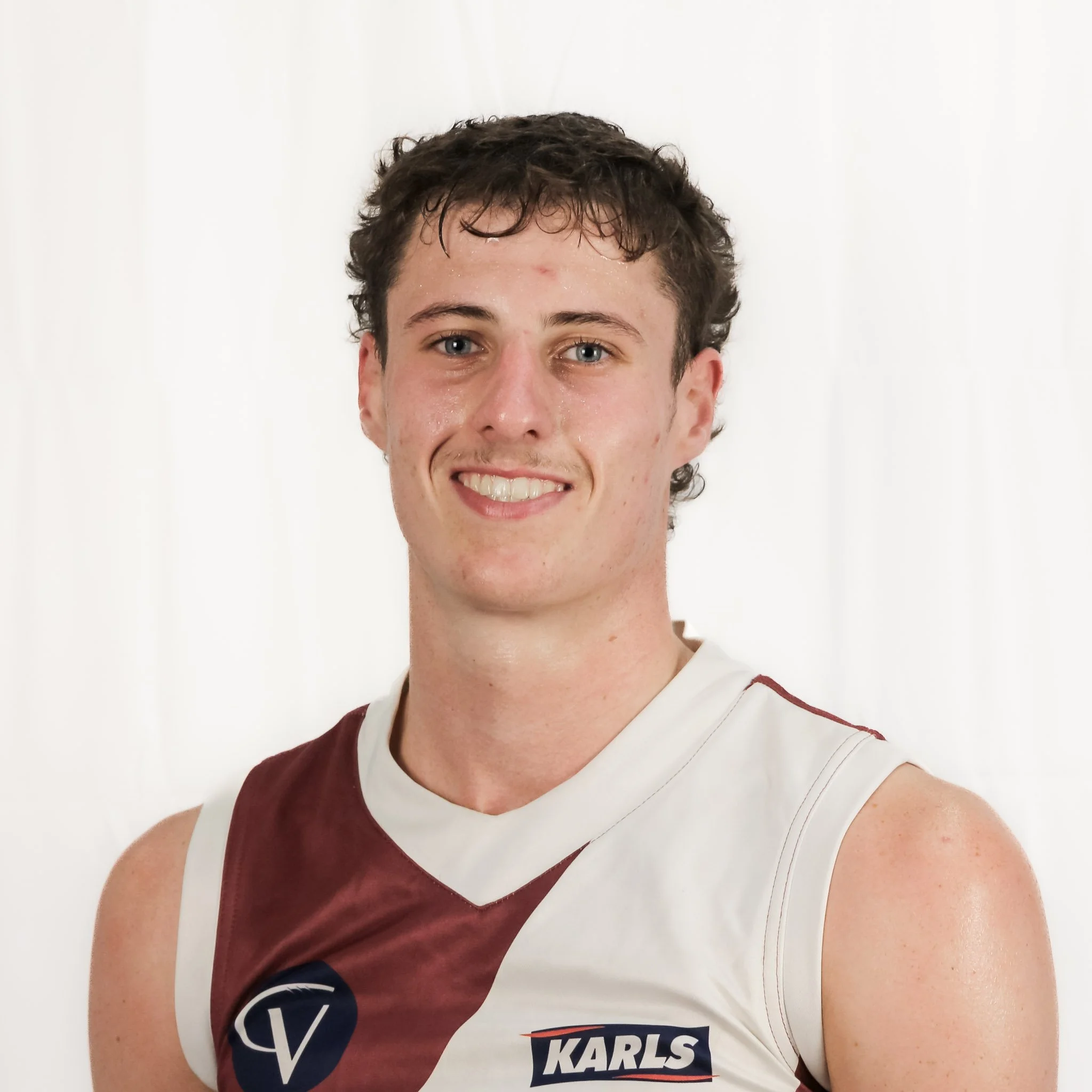 Portrait of a young man with short curly brown hair, blue eyes, and a smile, wearing a white and maroon sports jersey with sponsor logos, against a plain white background.