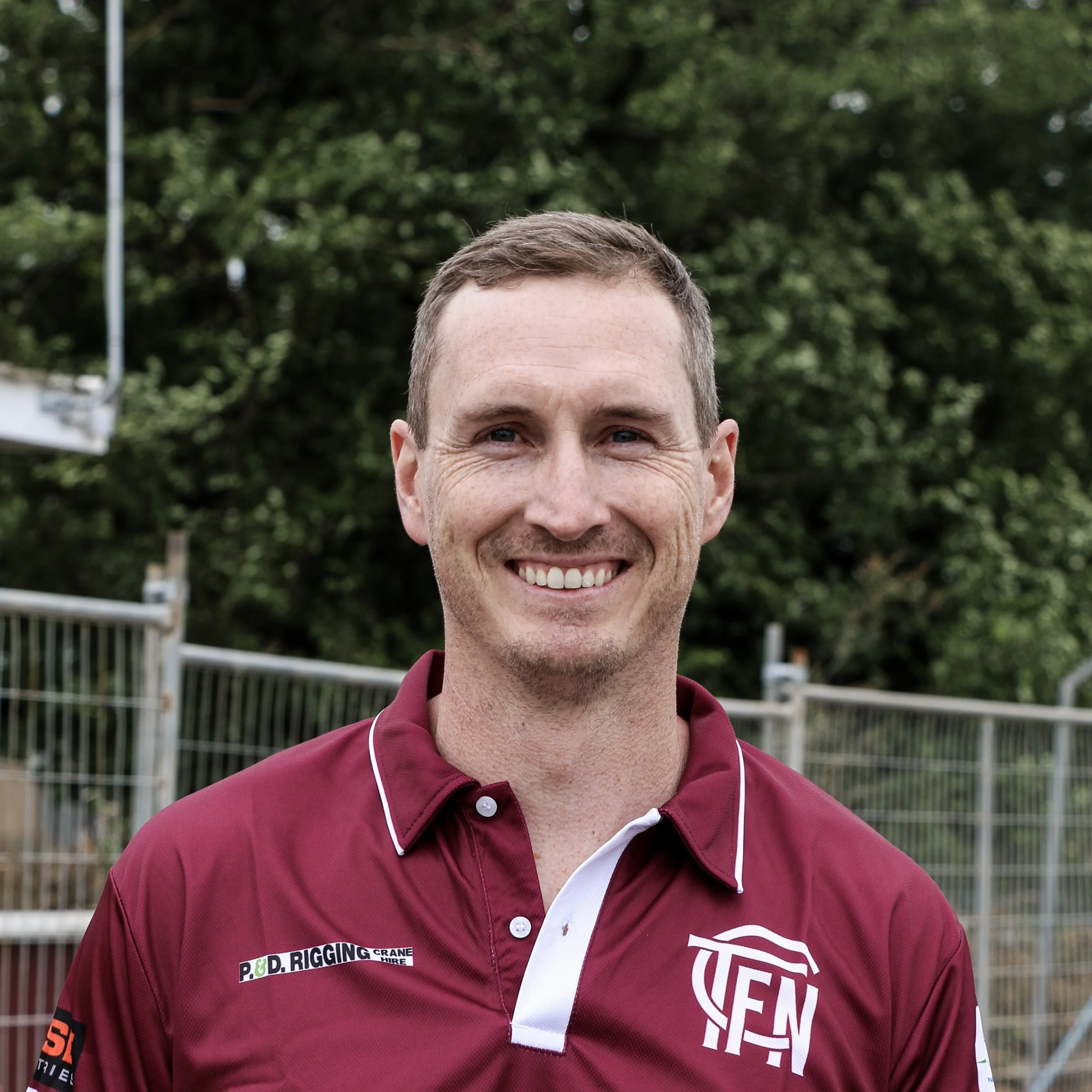 A smiling man in a maroon sports jersey standing outdoors with trees and a fence in the background.