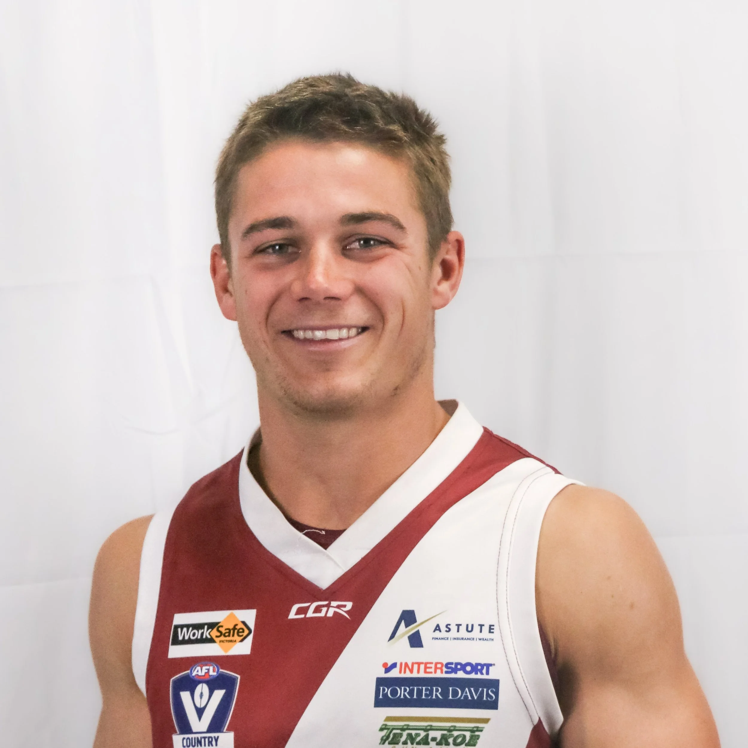 Close-up of a young man with short, light brown hair smiling, wearing a red and white sports jersey with multiple sponsor logos on it, against a plain white background.