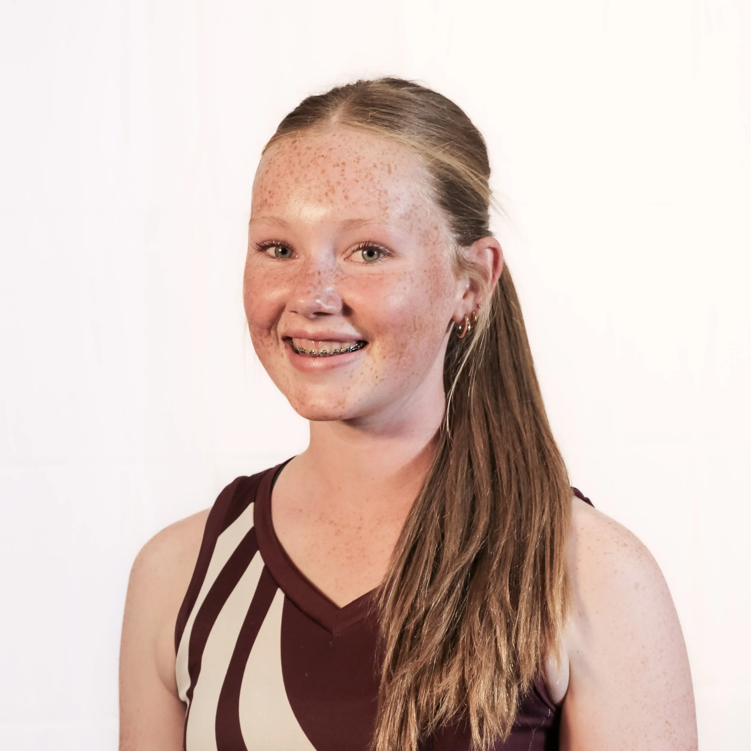 A young woman with long, straight, light brown hair and fair skin with freckles smiling at the camera. She is wearing a sleeveless top with maroon and white stripes on the shoulders and small hoop earrings. The background is plain white.