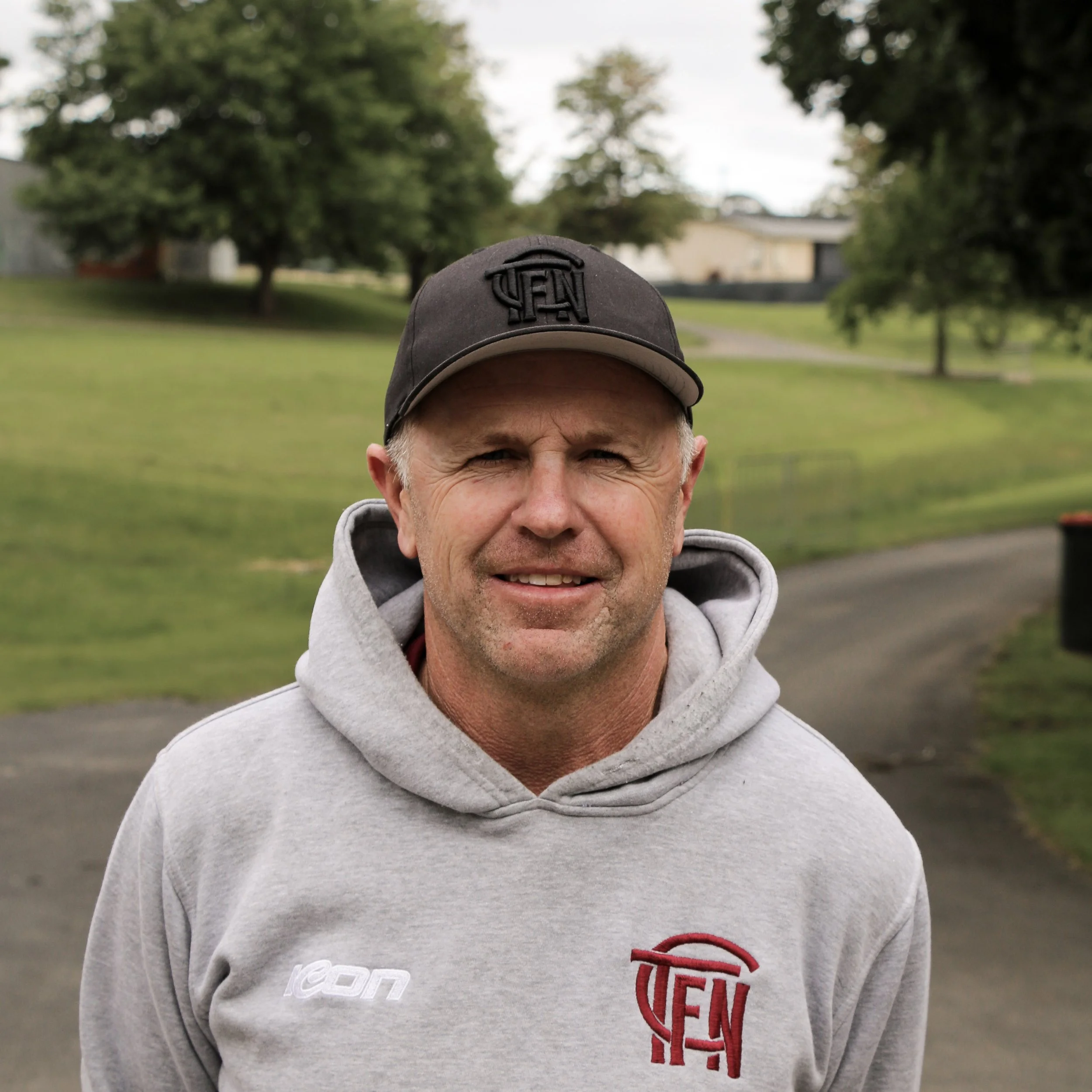 A man wearing a black baseball cap and gray hoodie with a red logo, standing outdoors on a paved path with green grass and trees in the background.