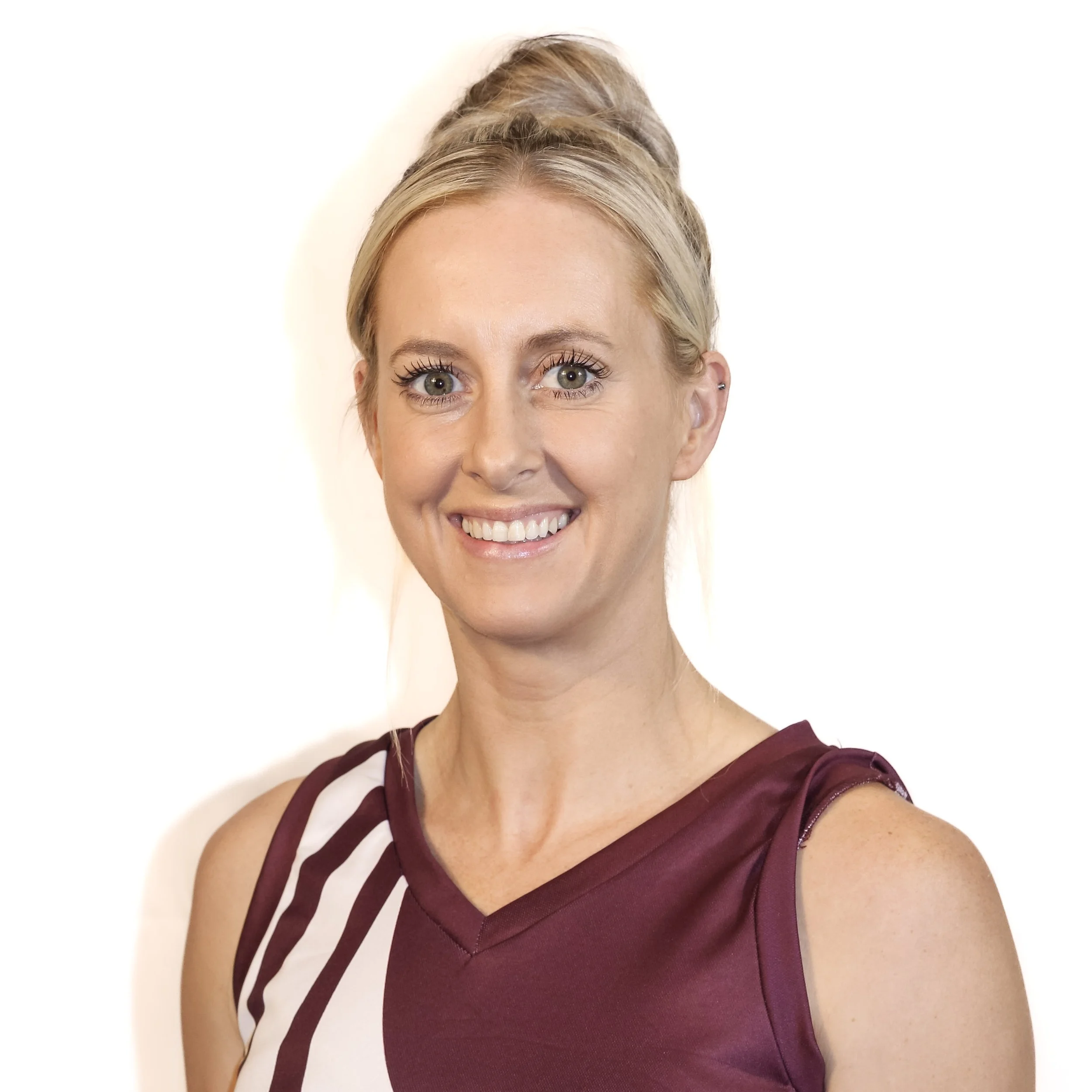Close-up of a smiling woman with blonde hair styled in an updo, wearing a sleeveless burgundy dress with cutout details, standing against a white background.