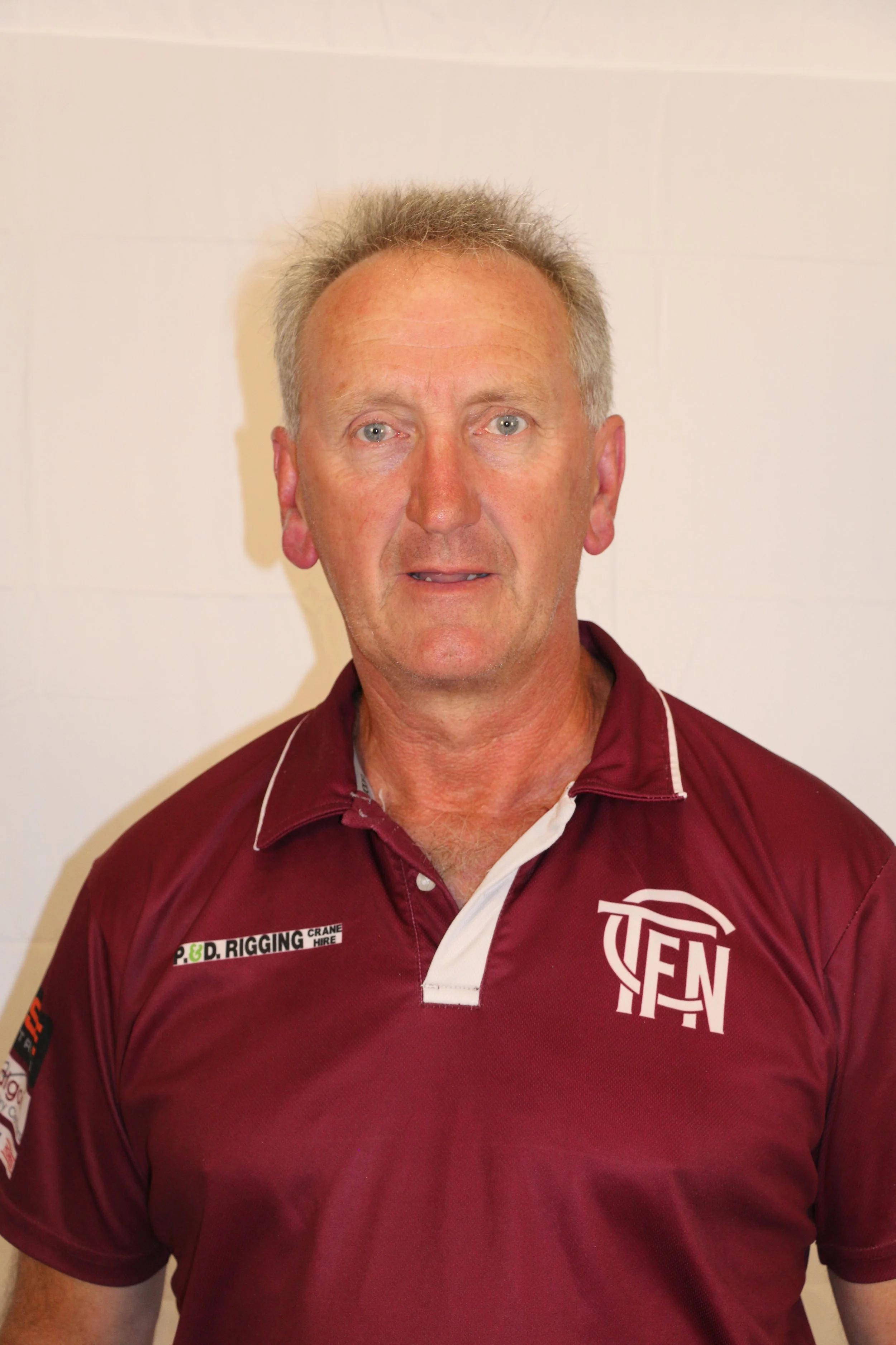 A man with light skin, short gray hair, and blue eyes is wearing a maroon collared shirt with sponsor patches and logos, standing against a plain white wall.