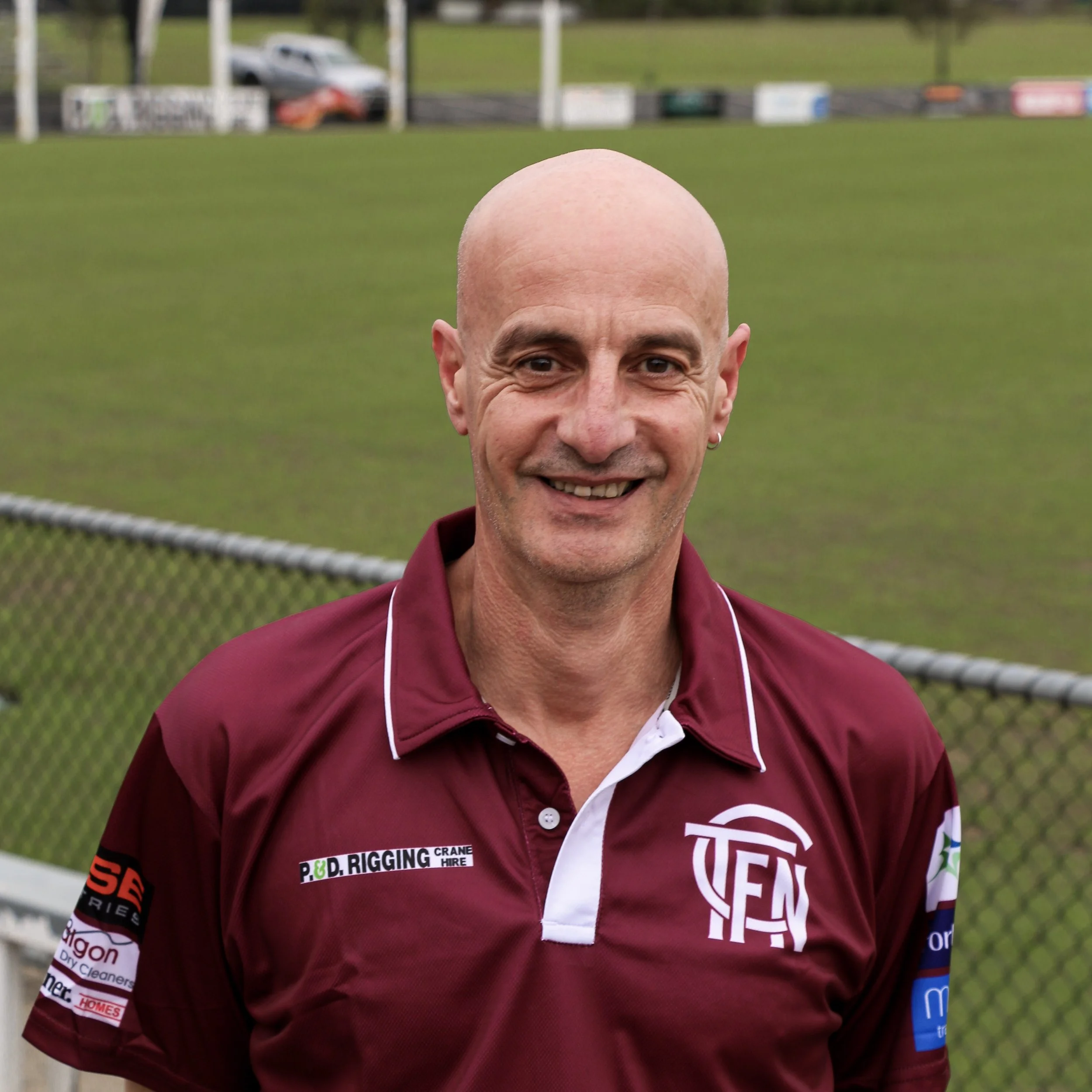 A smiling man with a shaved head, wearing a maroon sports shirt with white details, standing on a sports field with a fence and blurred vehicles in the background.