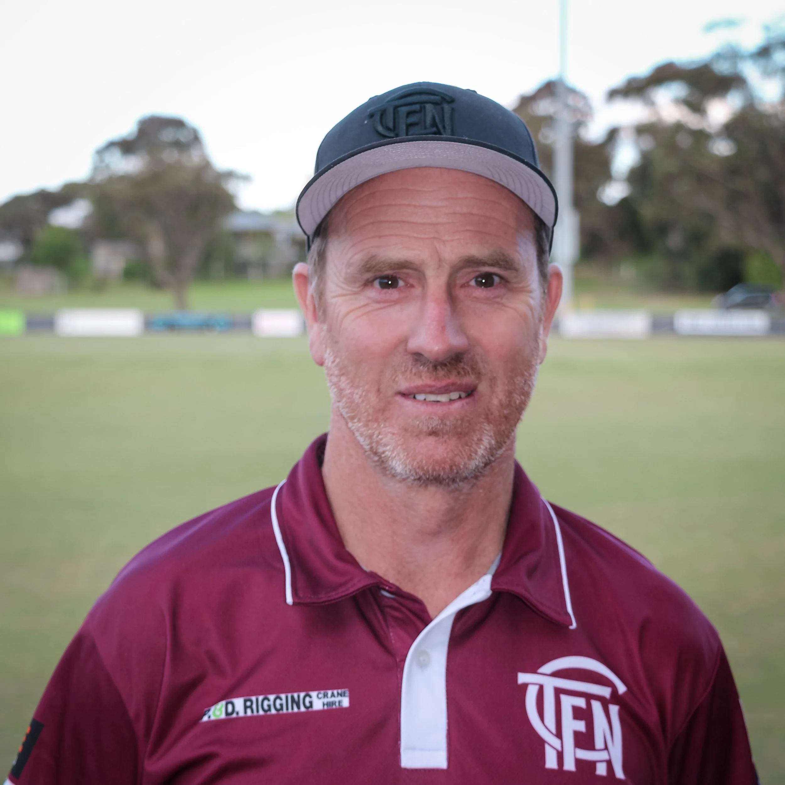 A middle-aged man in a maroon sports shirt and a black baseball cap, standing on a sports field with trees and a fence in the background.