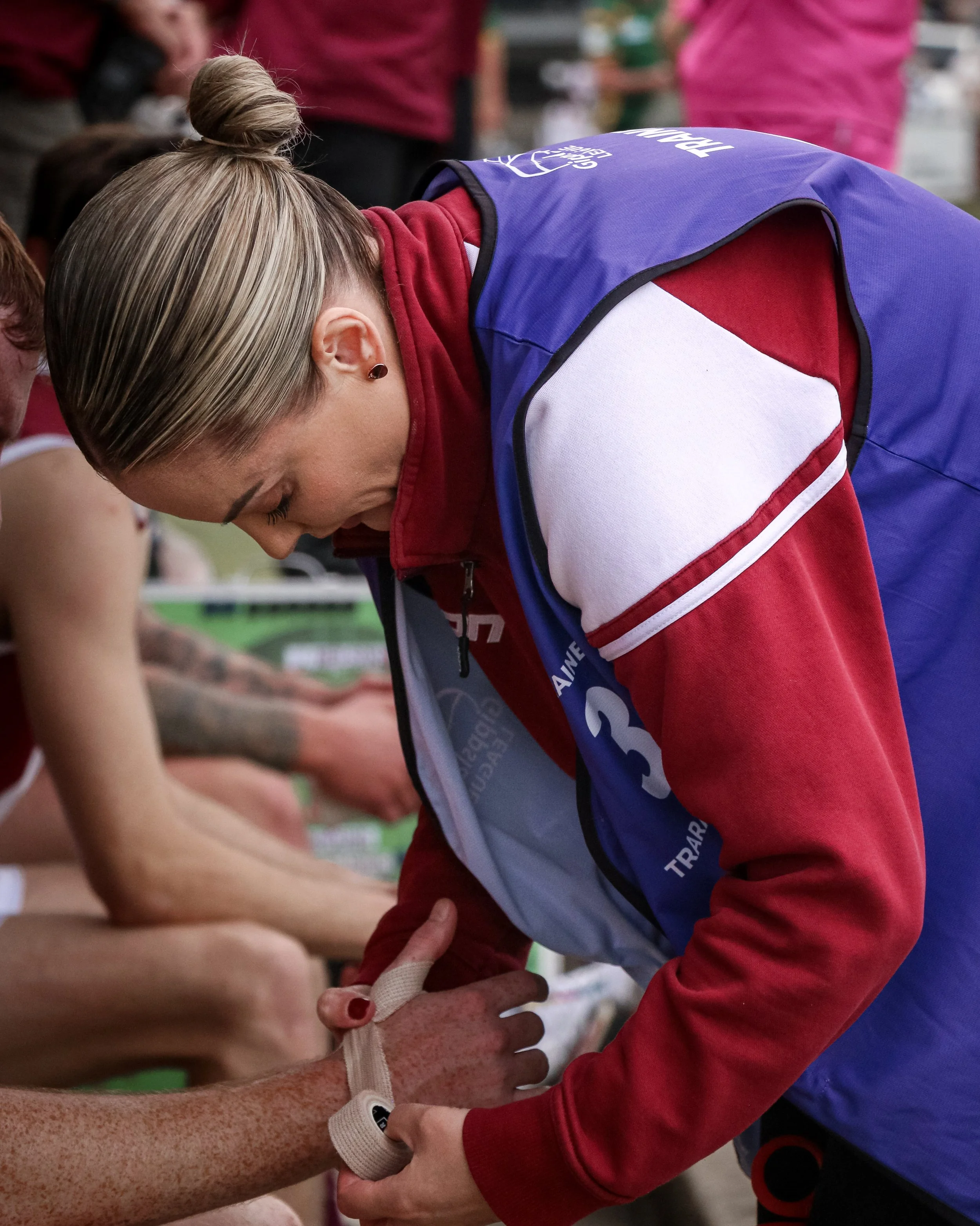 A woman with blonde hair tied in a bun is linking arms with a person wearing a university athletic jacket. The woman is wearing a red top and has earrings. The background shows other people, some in pink and red clothing, and a chain-link fence.