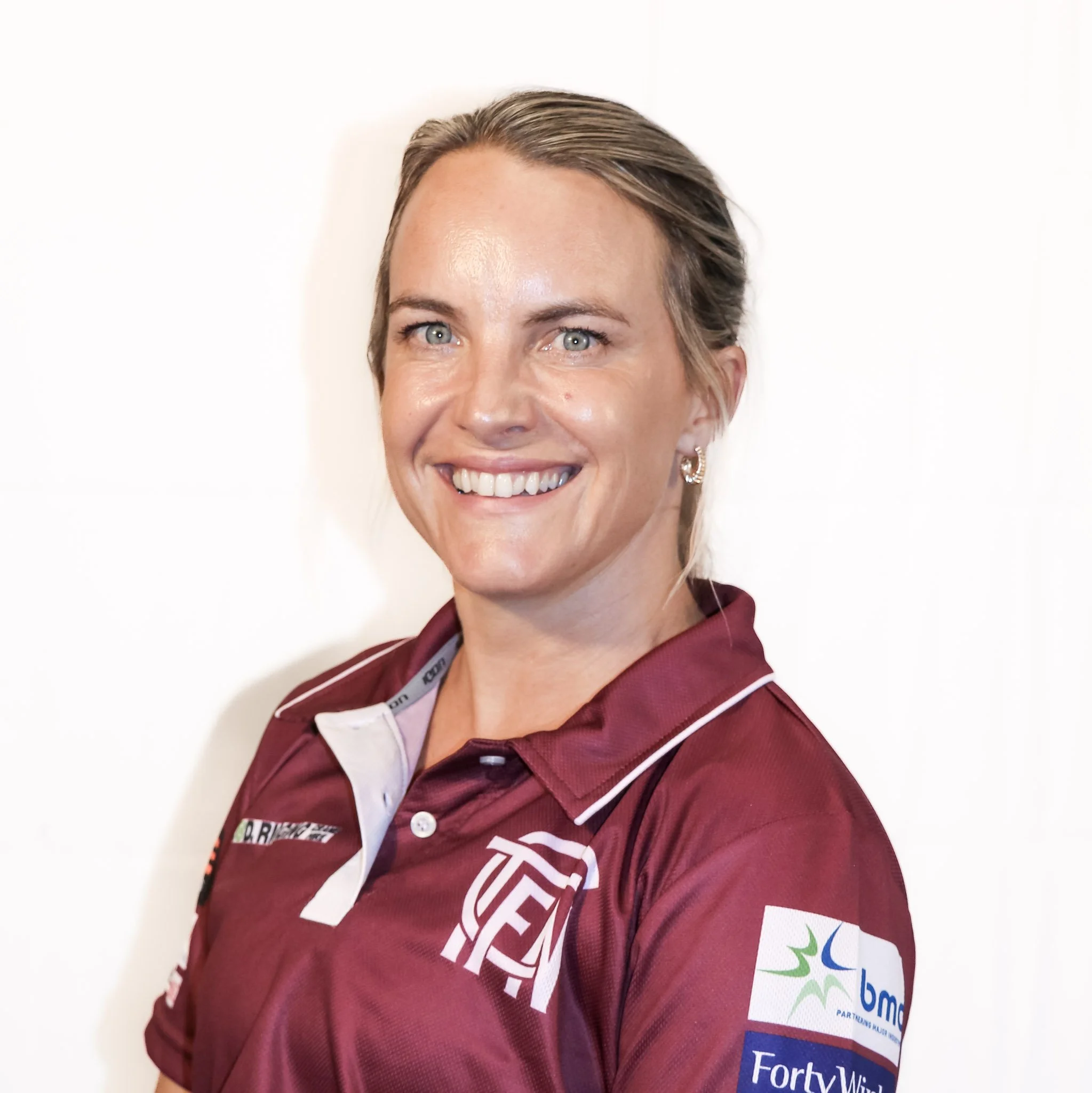 Close-up of a woman wearing a maroon shirt with network logos, smiling with short, light brown hair and blue eyes.
