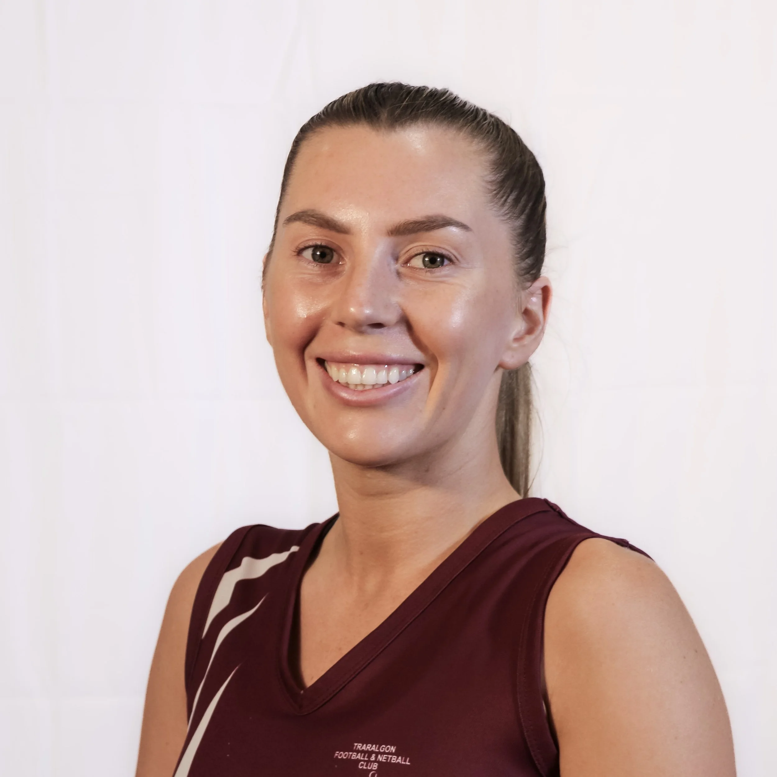 Close-up of a smiling woman with brown hair tied back, wearing a maroon sports top with white and black stripes, against a white background.