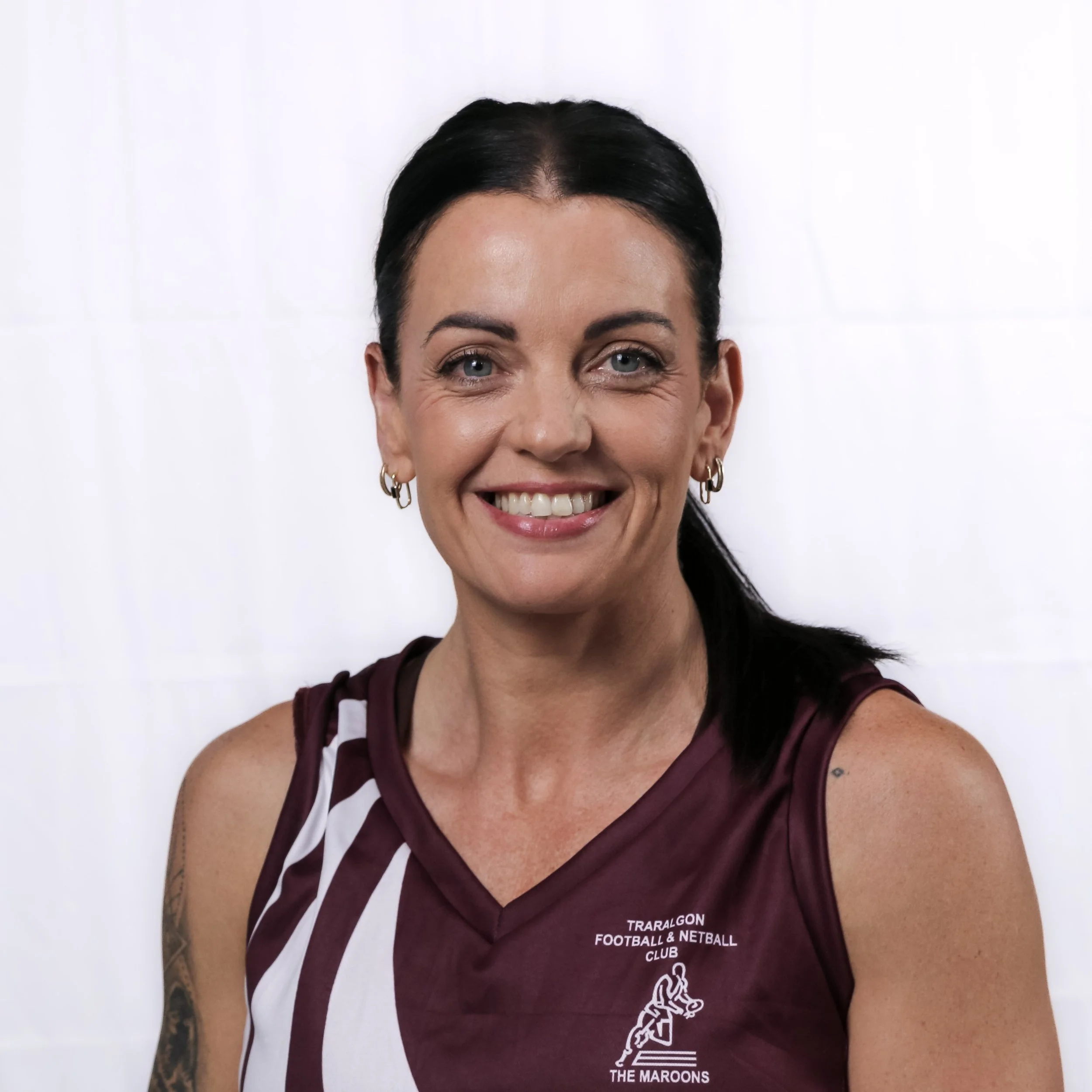 A woman with dark hair and blue eyes smiling, wearing a maroon sports shirt with white text and logos, against a plain white background.