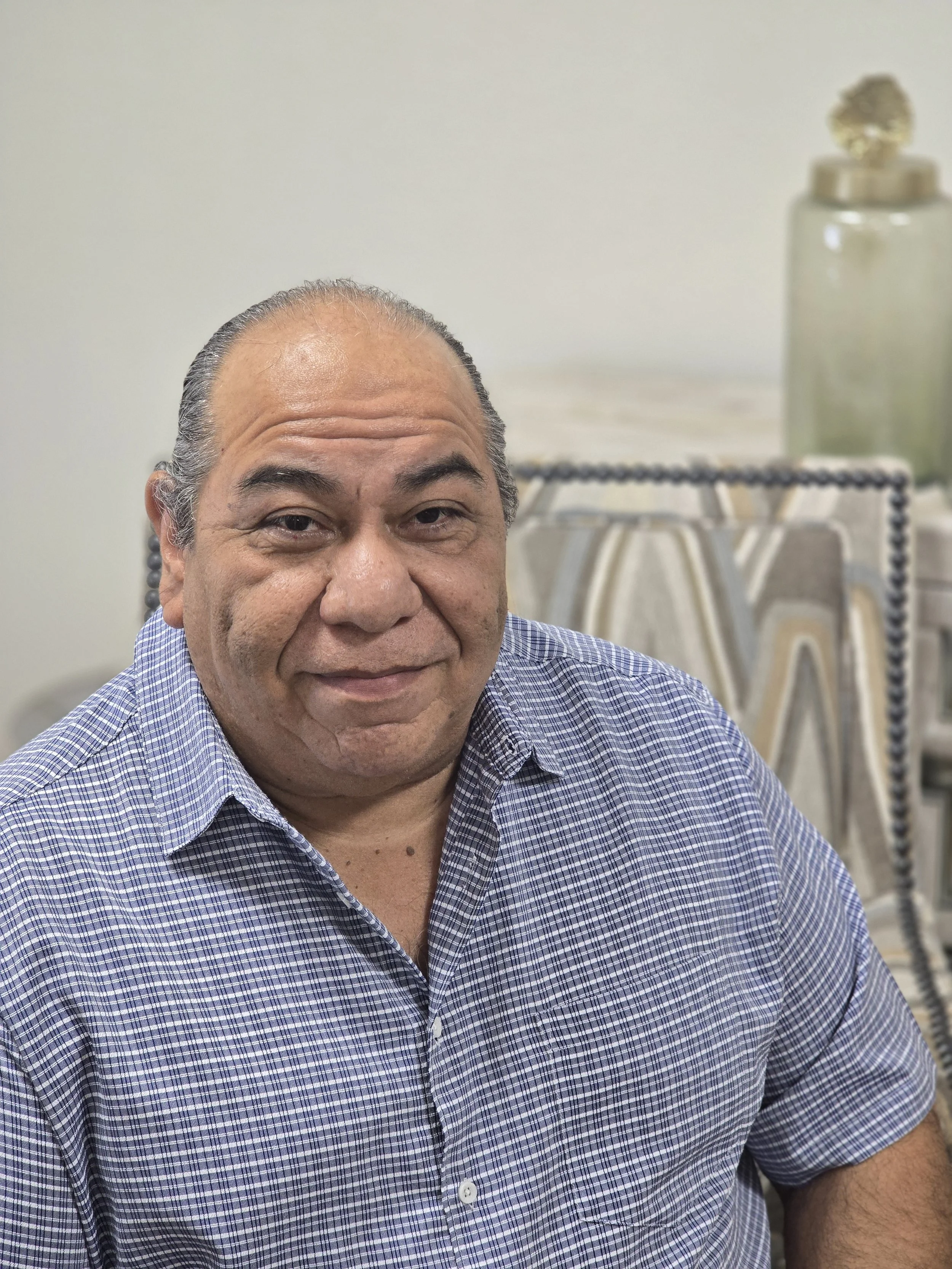 A middle-aged man with dark hair, partially gray, slicked back, wearing a checkered blue shirt, sitting indoors with a neutral expression. In the background, there is a table with a decorative vase and a patterned chair.