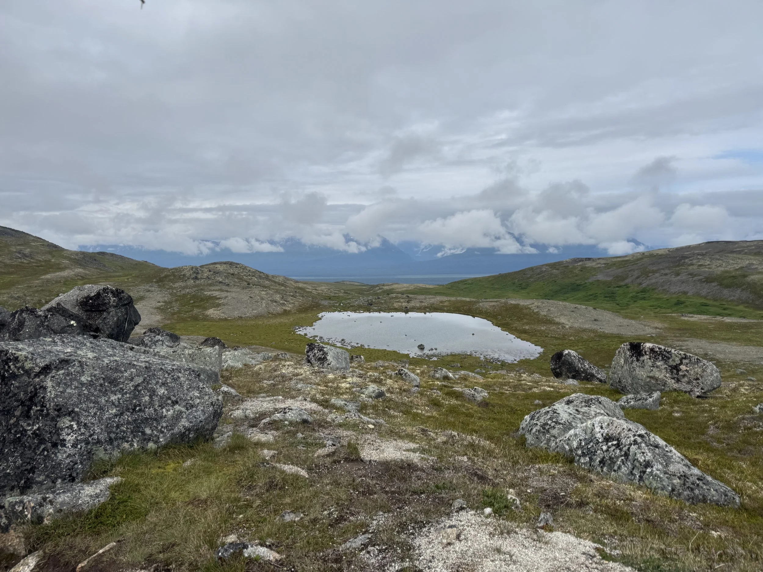 A landscape view of mountains covered with snow in the distance under a cloudy sky, with a small lake and rocks in the foreground.