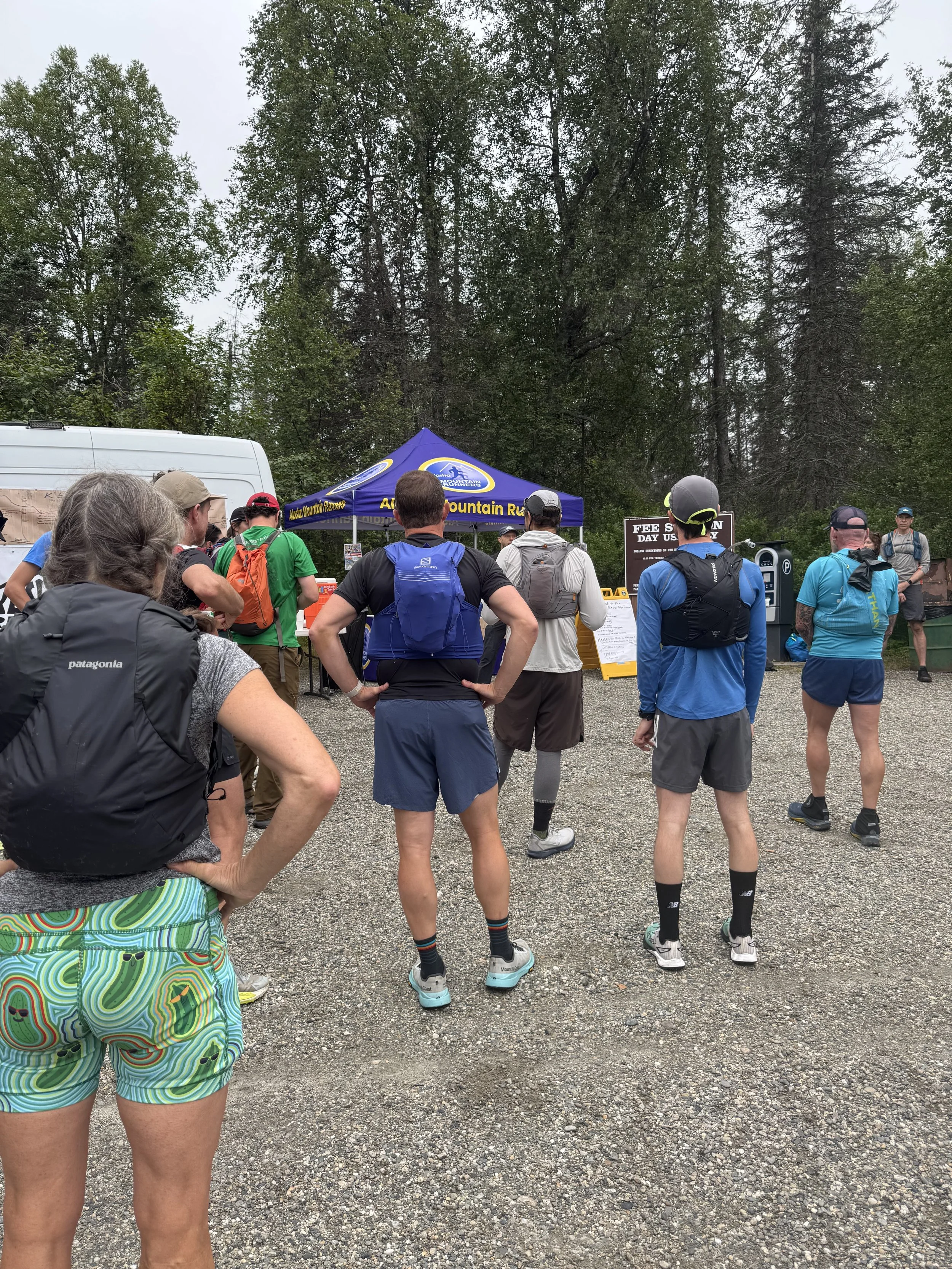 Group of people in athletic gear gathered outdoors near a blue canopy with a sign, in a wooded area on a cloudy day.