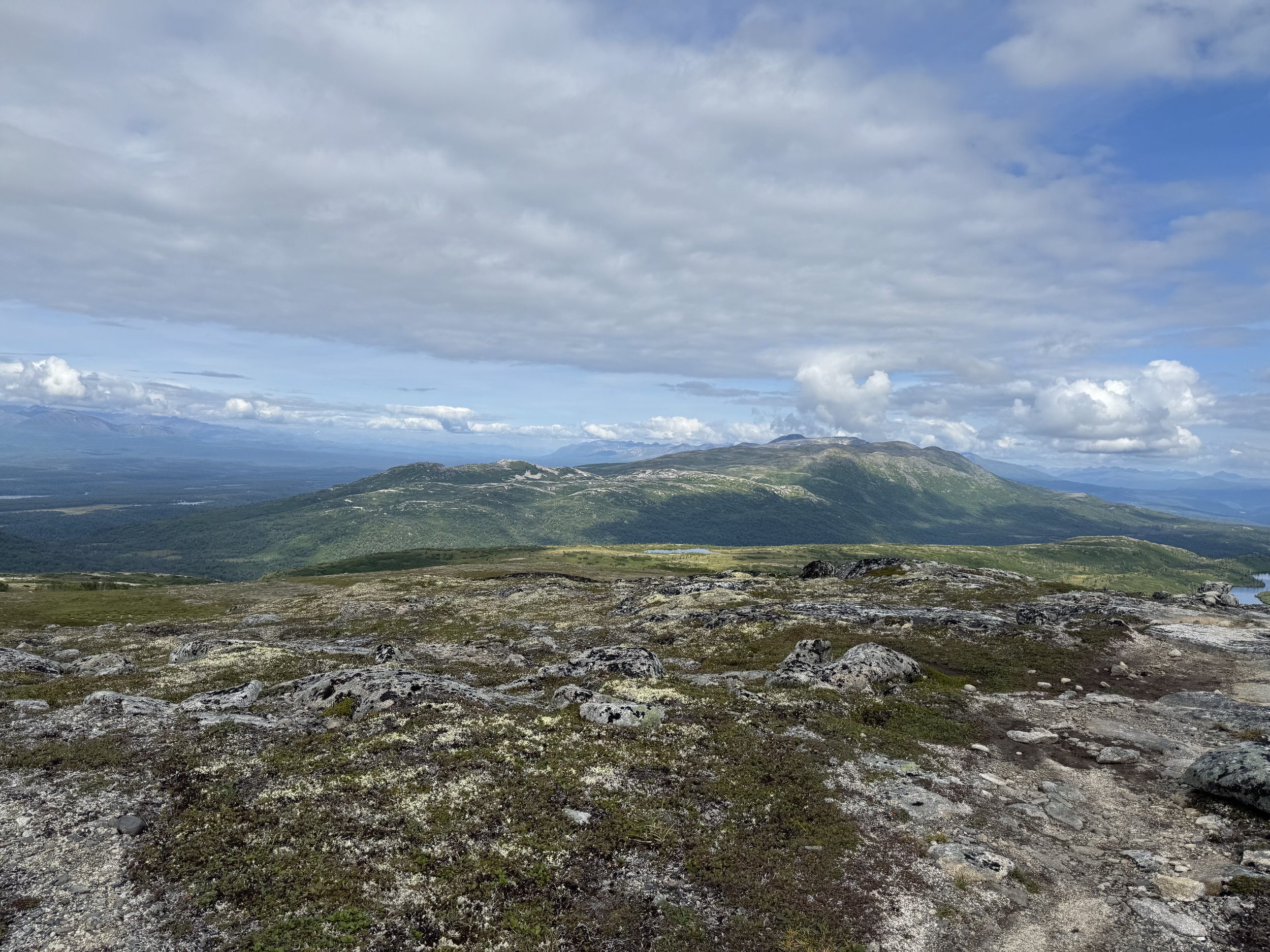 A scenic view of rolling green hills and mountains under a cloudy sky, with patches of blue sky visible.