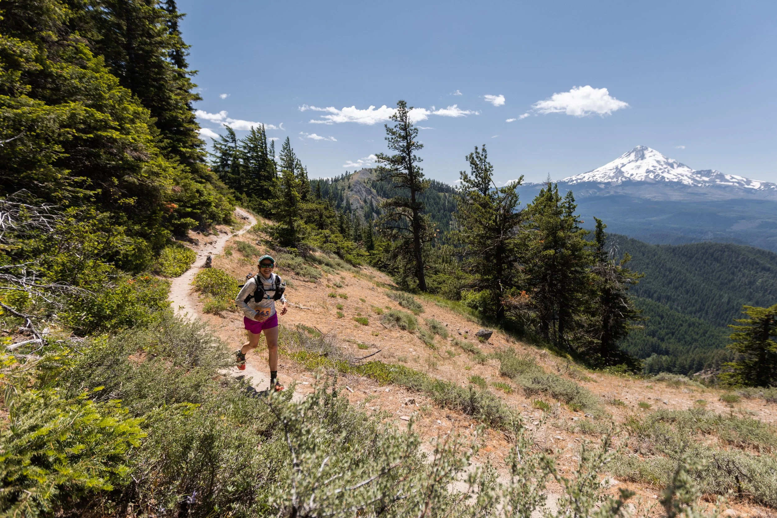 A woman trail running on a dirt path through a forest with pine trees, a mountain with snow in the background, and a clear blue sky with some white clouds. Wy'east Wonder