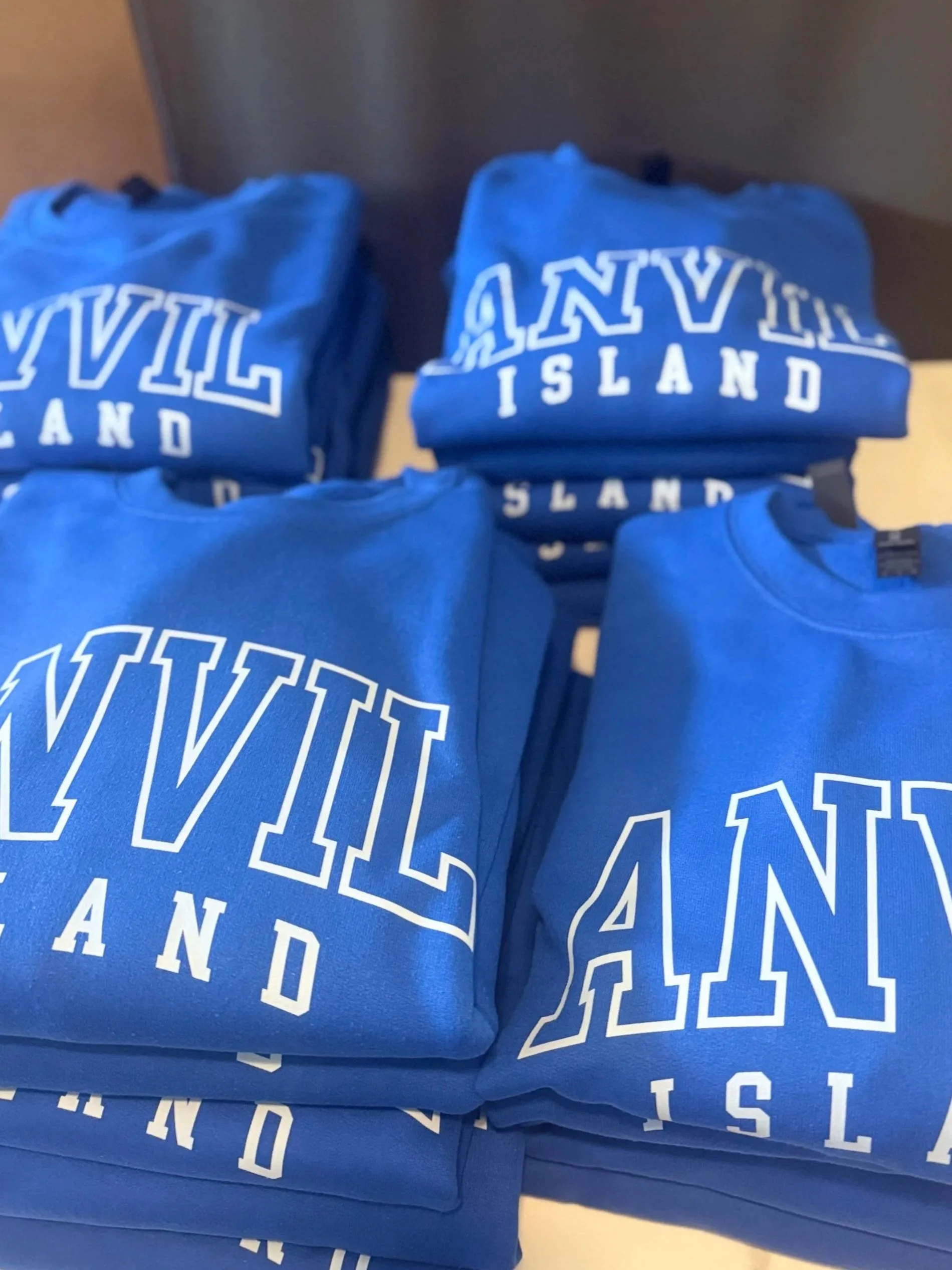 Multiple blue sweatshirts with white text reading "VANVIL ISLAND" stacked on a table.