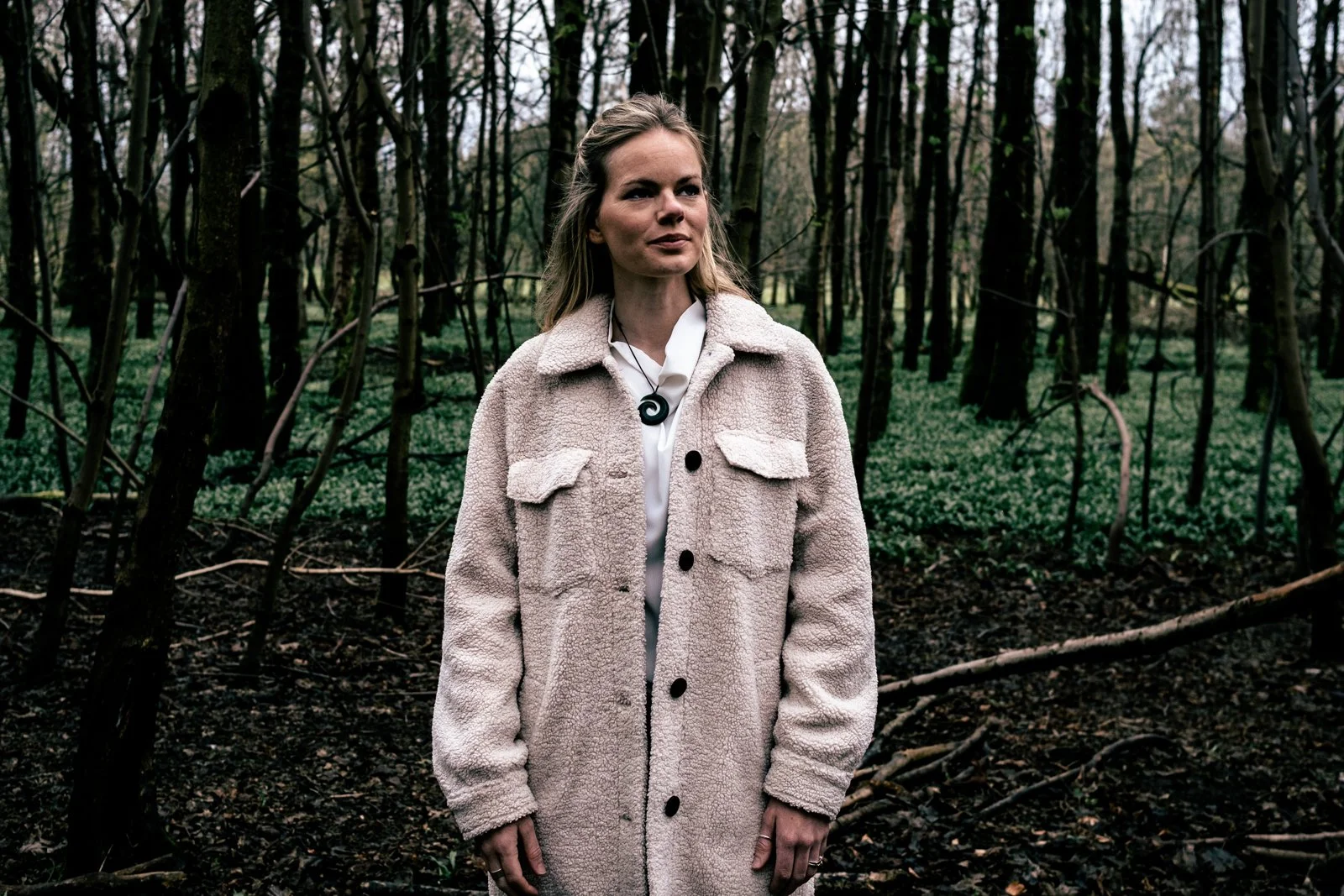 Woman in a light coat standing in a forest, surrounded by trees and greenery.