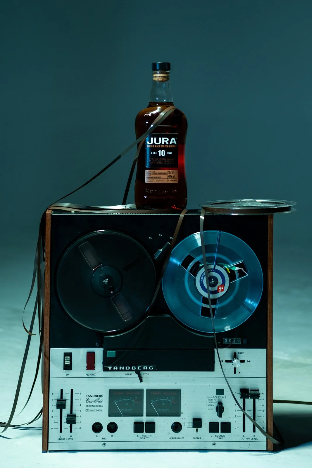 A vintage reel-to-reel tape recorder with a tape running across the reels, sitting on a surface. On top of the recorder is a bottle of Jura 10-year-old single malt scotch whisky. The setting is dimly lit, creating a moody atmosphere.