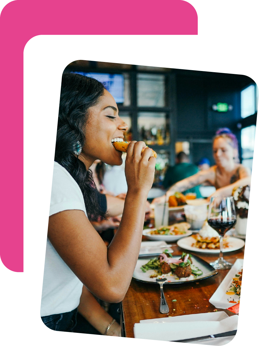 A woman with dark hair and earrings eating a piece of fried chicken in a restaurant or bar with other people dining in the background, along with plates of food, a glass of red wine, and a lively atmosphere.