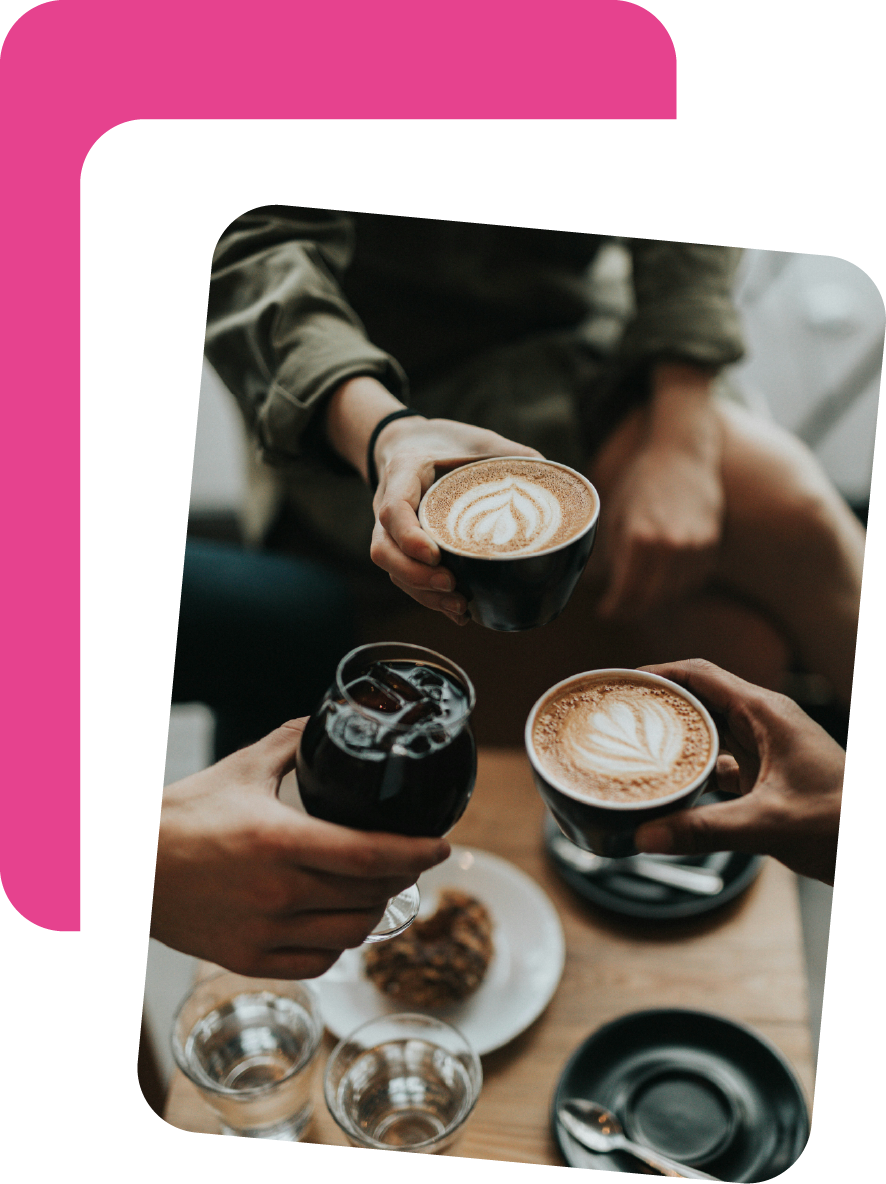 Three people holding cups of coffee with latte art, a glass of soda, and a plate with a pastry on a wooden table.