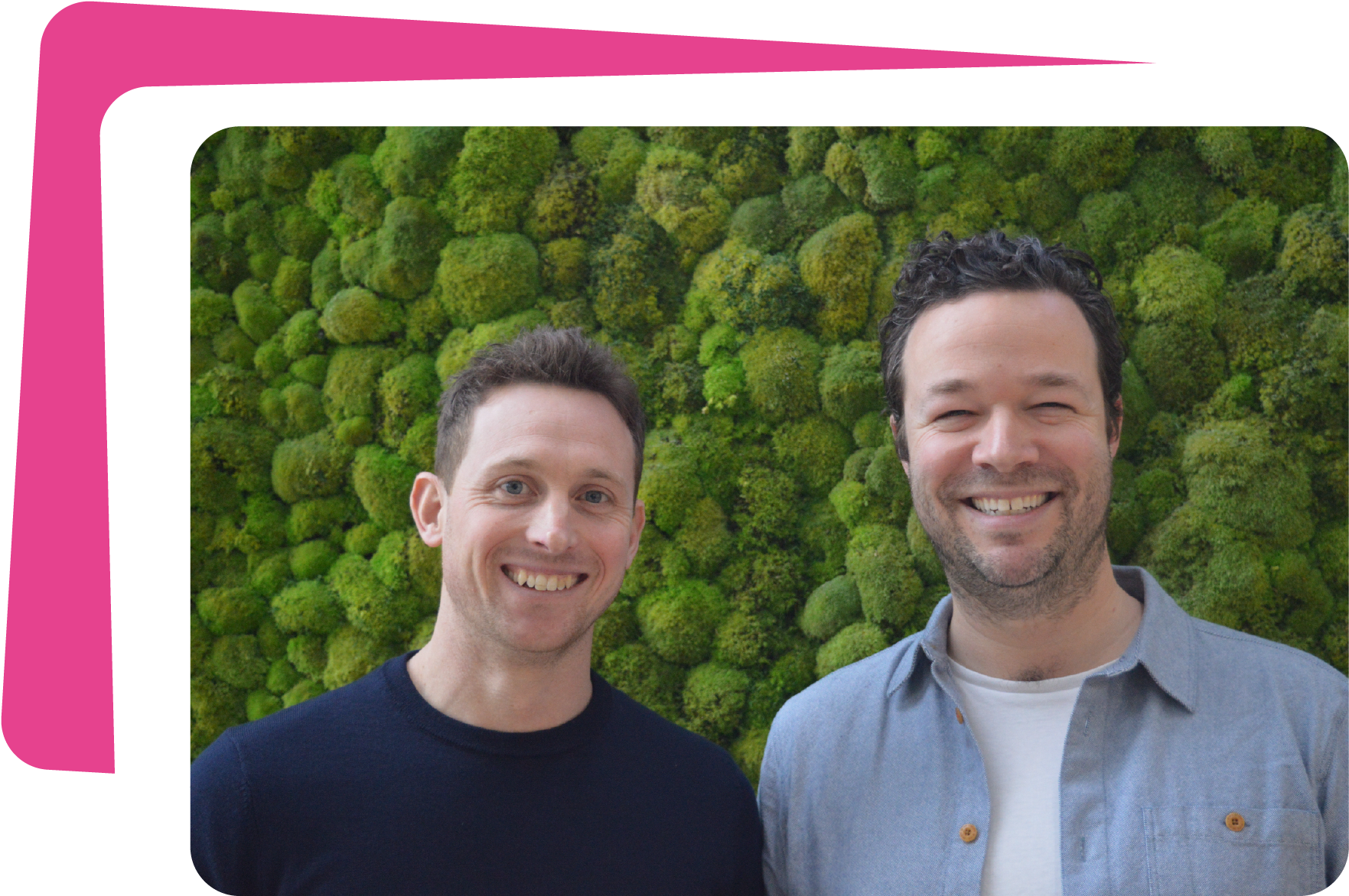 Two smiling men standing outdoors in front of a green moss-covered wall.