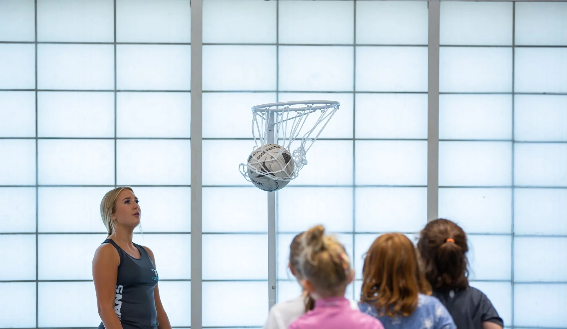 A woman in athletic clothing appears to be taking a netball shot in an indoor court, with three children watching in the foreground.
