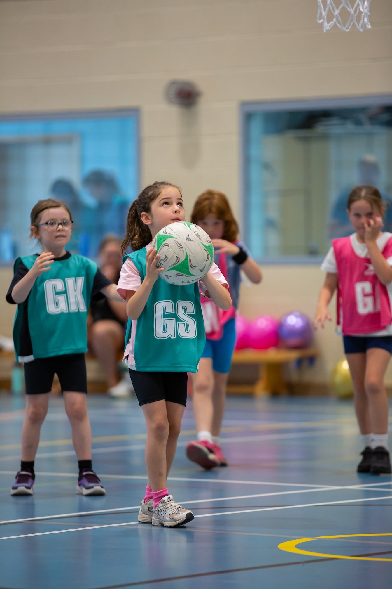 Young girls playing netball in an indoor sports hall, with one girl in the center holding a netball, preparing to shoot, while others observe around her.