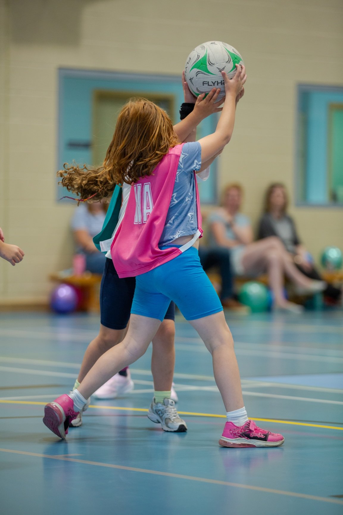 Young girl with red hair in a pink vest and blue shorts playing indoor junior netball, blocking the ball at the net, with spectators watching in the background.