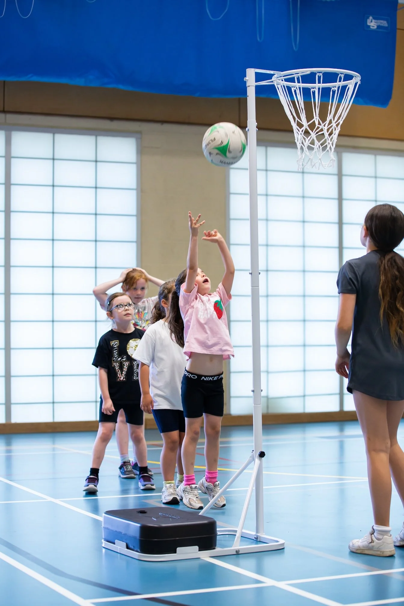 Children playing basketball in an indoor gymnasium, with one girl shooting the ball towards the hoop and others standing in line.