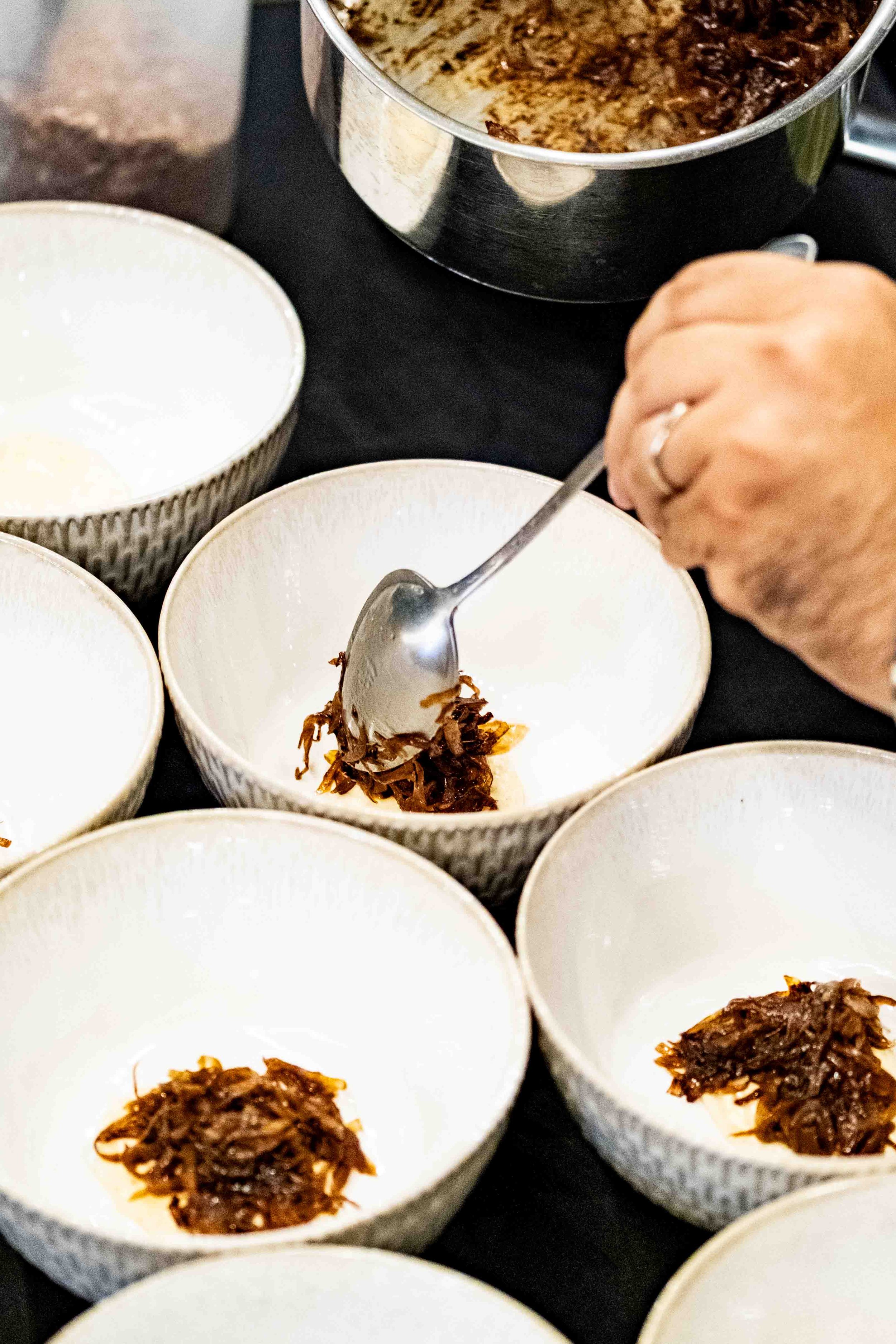 Person serving shredded beef or pulled meat into small white bowls using a spoon, with a large pot of cooked meat in the background.