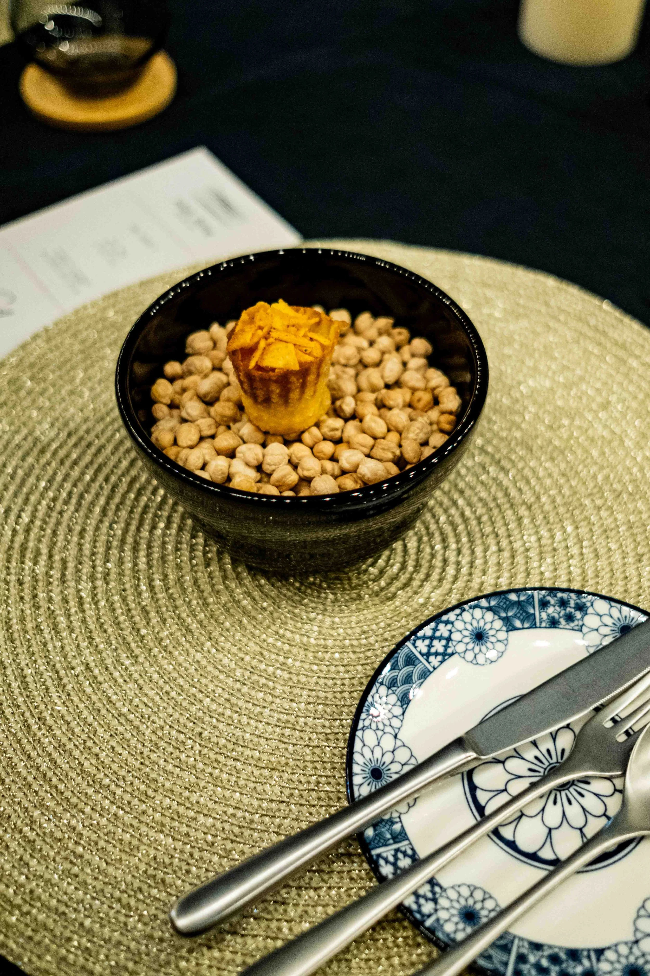 A small black bowl filled with round snack foods, topped with a small muffin, placed on a gold-colored placemat on a table. Next to the bowl is a blue and white floral patterned plate with a fork, knife, and spoon on it.