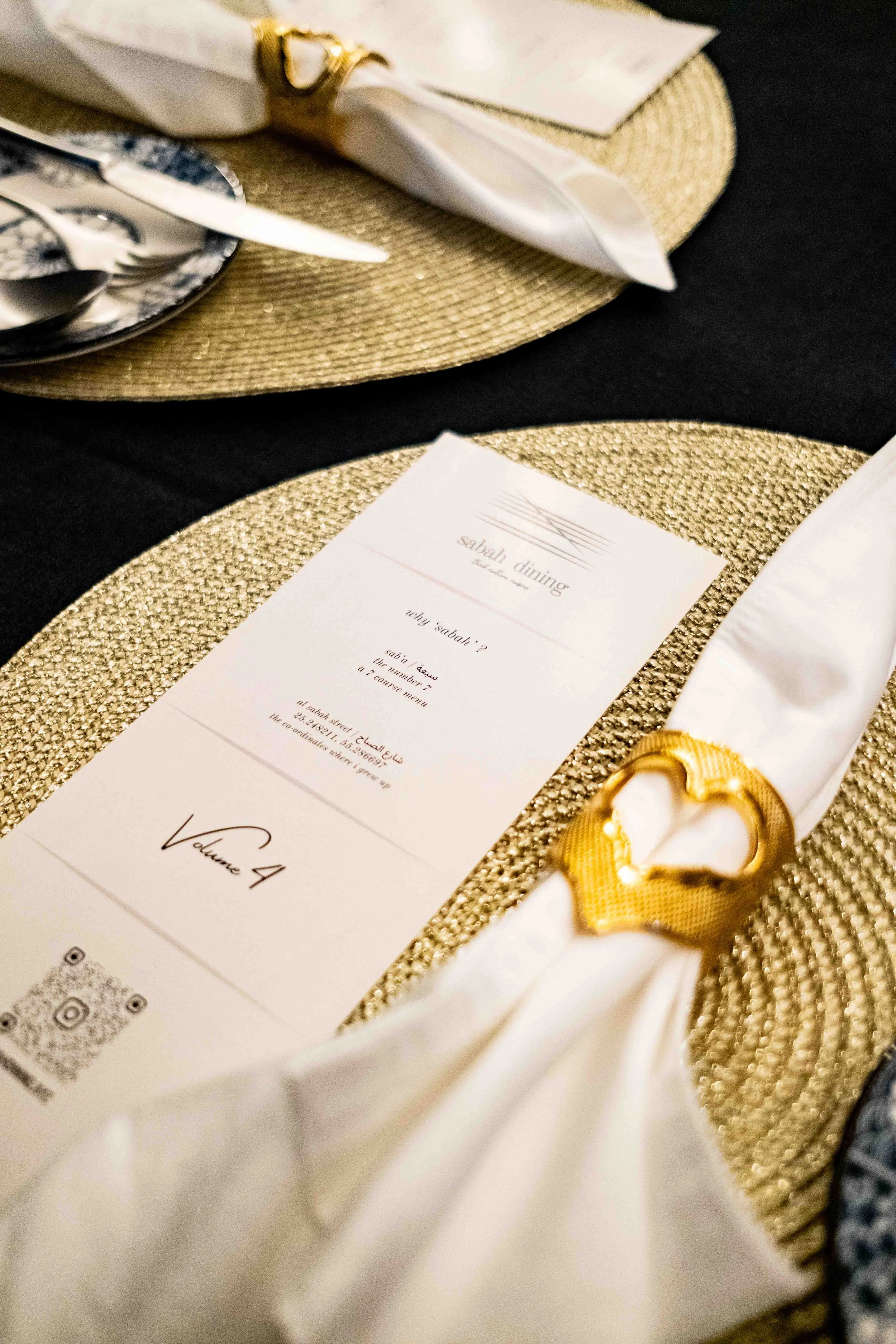Menu card on a gold placemat, decorated with a white napkin held by a gold napkin ring, set on a black table with a matching gold placemat.