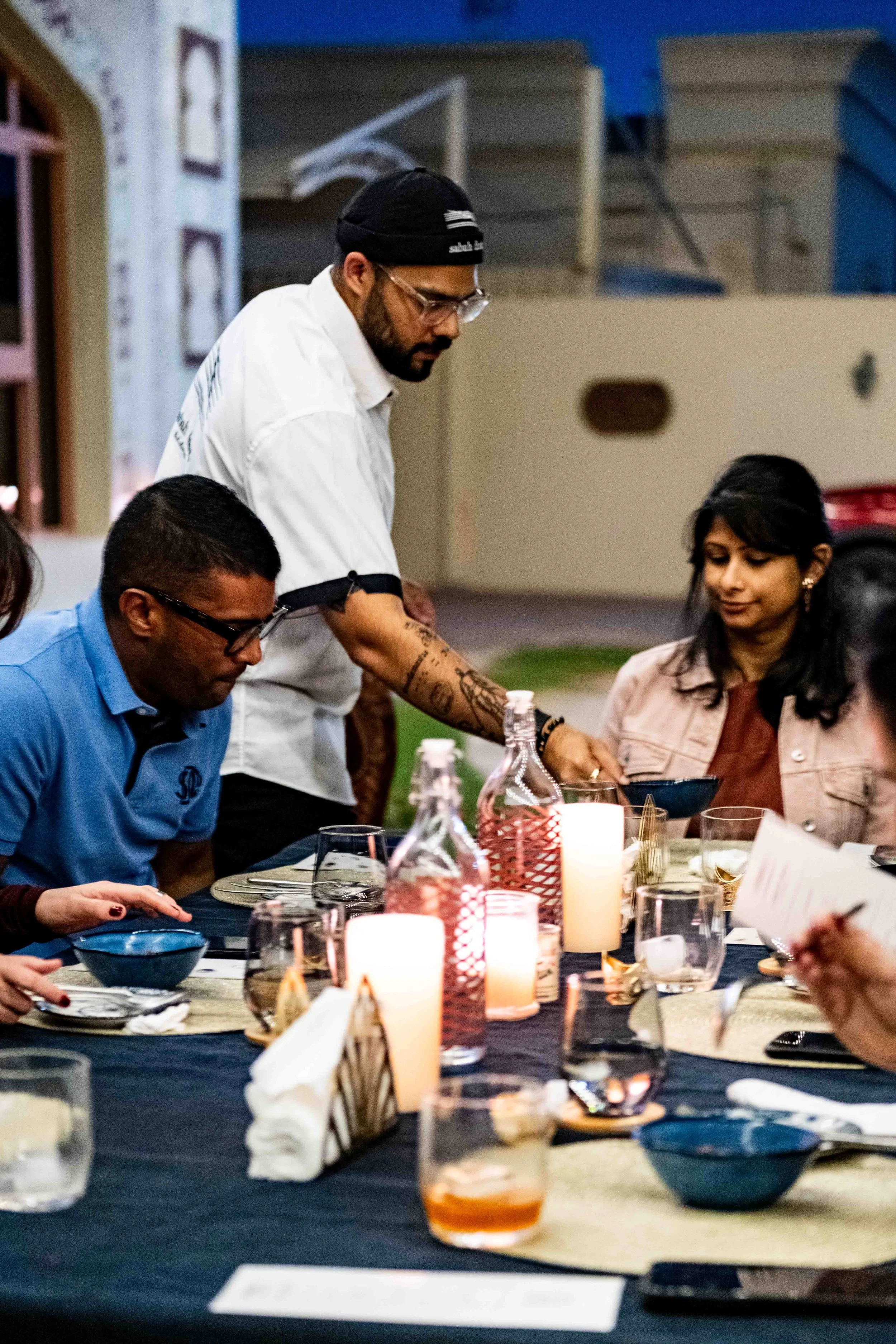 A man serving food at a dinner party with people seated at a table with candles and drinks outdoors.