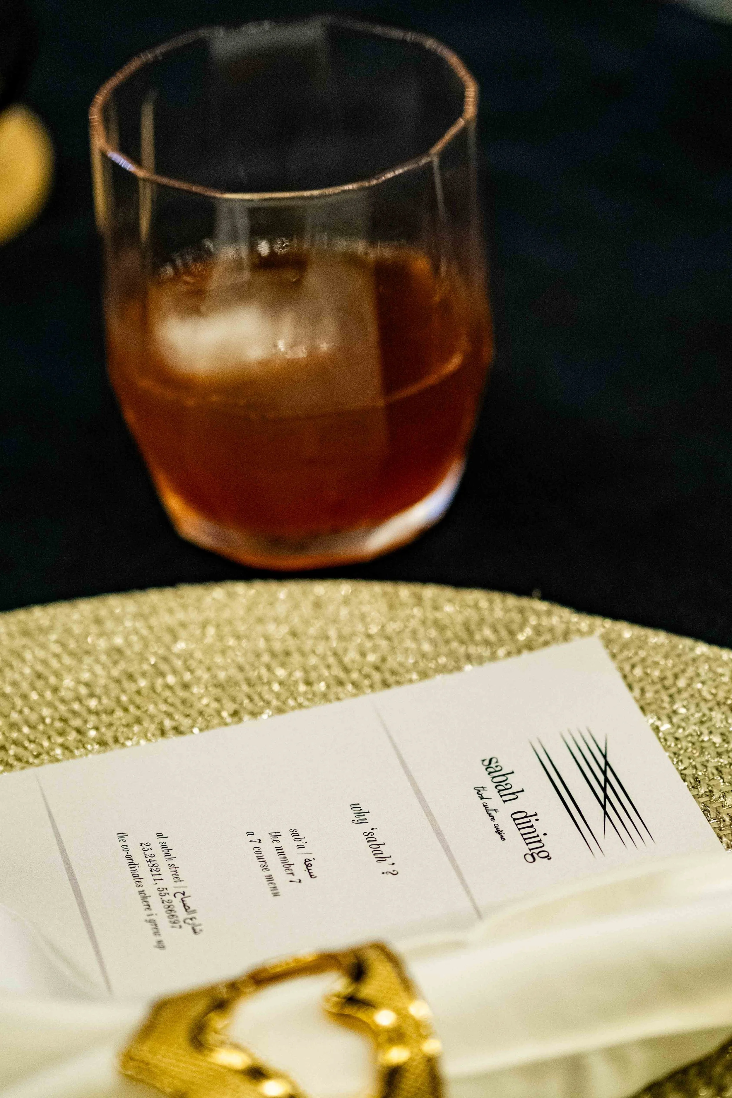 A glass of dark whiskey with a large ice cube, placed on a black surface, next to a silver glittery clutch purse and a white open book or menu with the title 'Sabad Dinner.'
