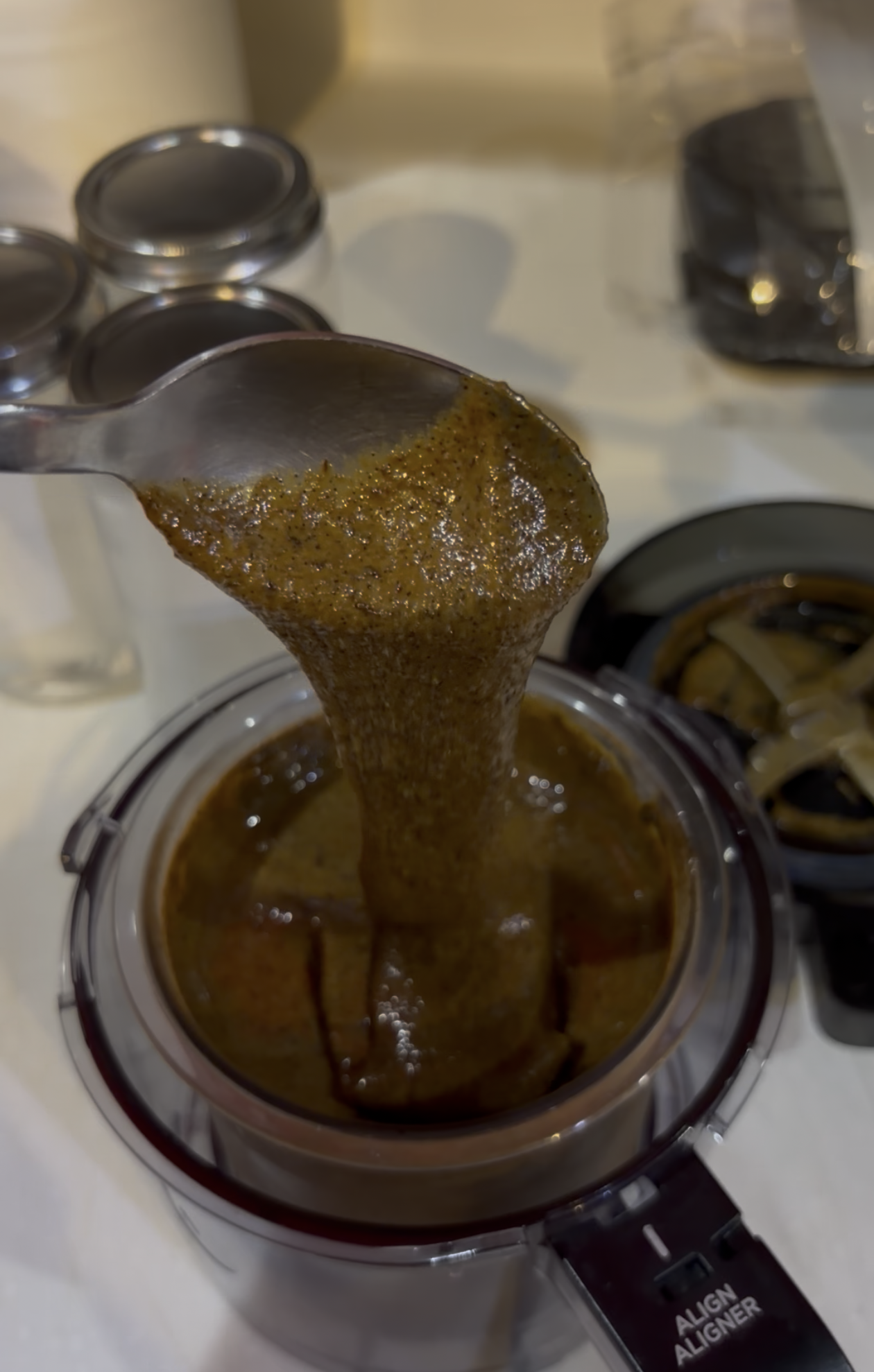 A spoon lifting a thick, brownish-green sauce or puree from a blender with ingredients and kitchen items in the background.