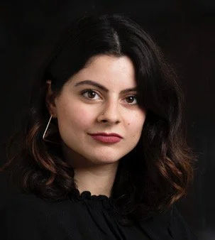 Portrait of a woman with dark wavy hair, wearing a black top, against a dark background.