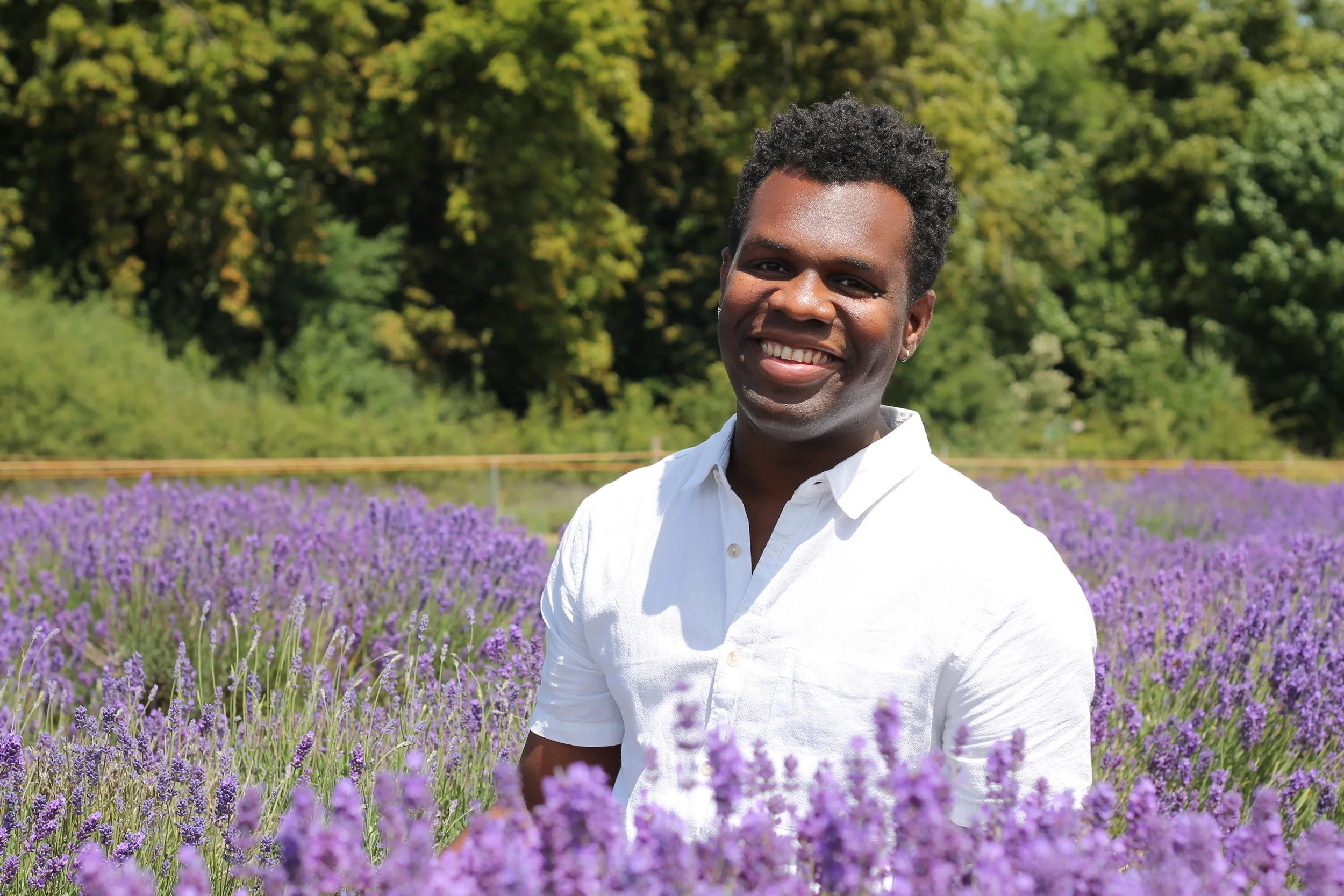 A smiling man standing in a lavender field with green trees in the background.