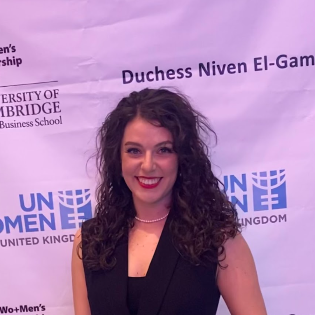 Woman with curly hair smiling in front of a UN Women UK backdrop.