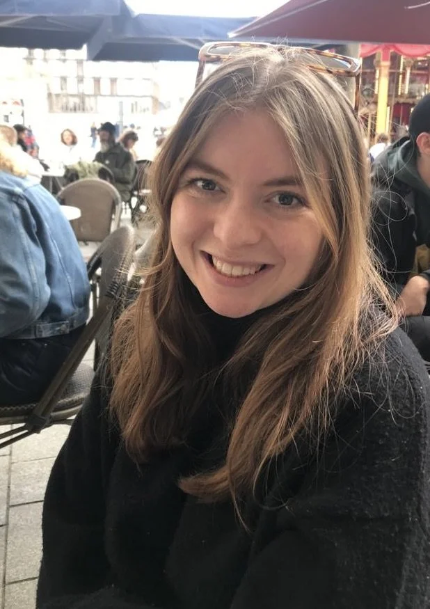 A young woman with long light brown hair, wearing sunglasses on her head, smiling at an outdoor cafe with people seated at tables under umbrellas.