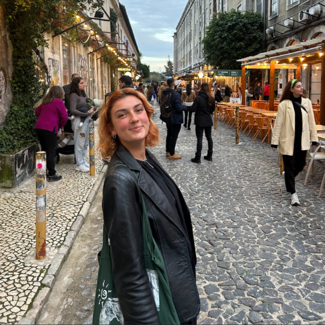 A young woman with curly red hair smiling and looking at the camera on a cobblestone street in a lively outdoor area with shops and cafes. Other people are walking and chatting, with some sitting at outdoor tables. The scene is during evening with string lights illuminating the area.