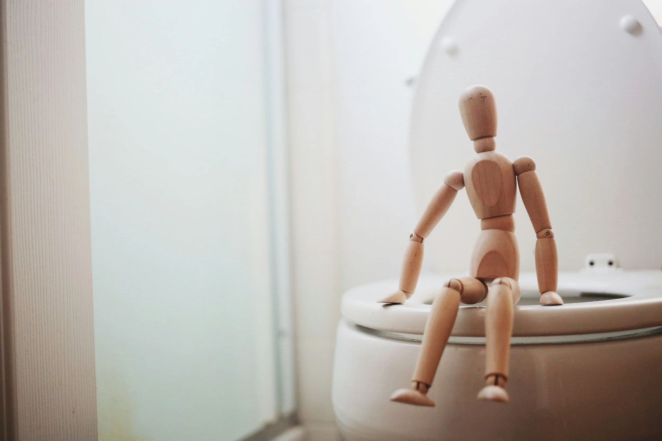 A wooden artist mannequin sitting on the edge of a toilet in a bathroom.