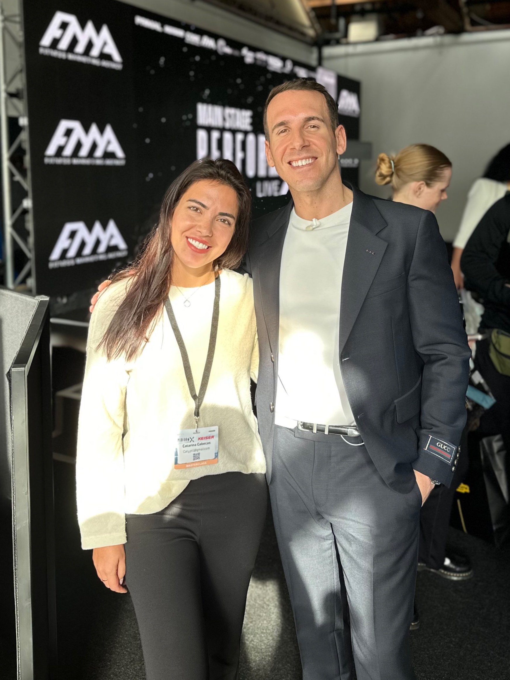 Two people smiling and posing for a photo at an event, with a backdrop displaying logos and the words "Main Stage Perform." One person is wearing a name badge.