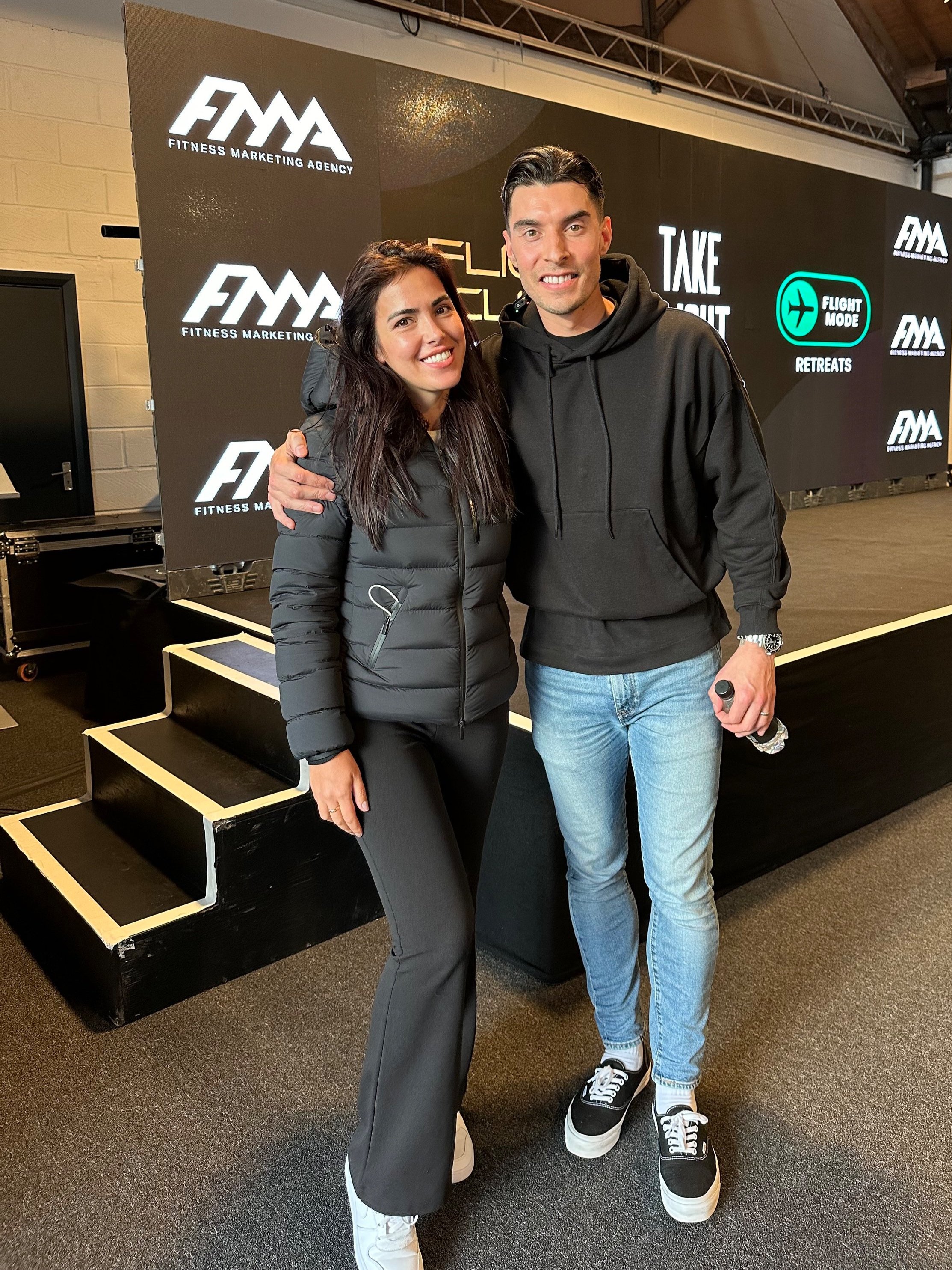 Two people posing in front of a fitness marketing agency backdrop at an event, with branded logos and displays in the background. One is wearing a black hoodie and jeans, the other a black jacket and pants.
