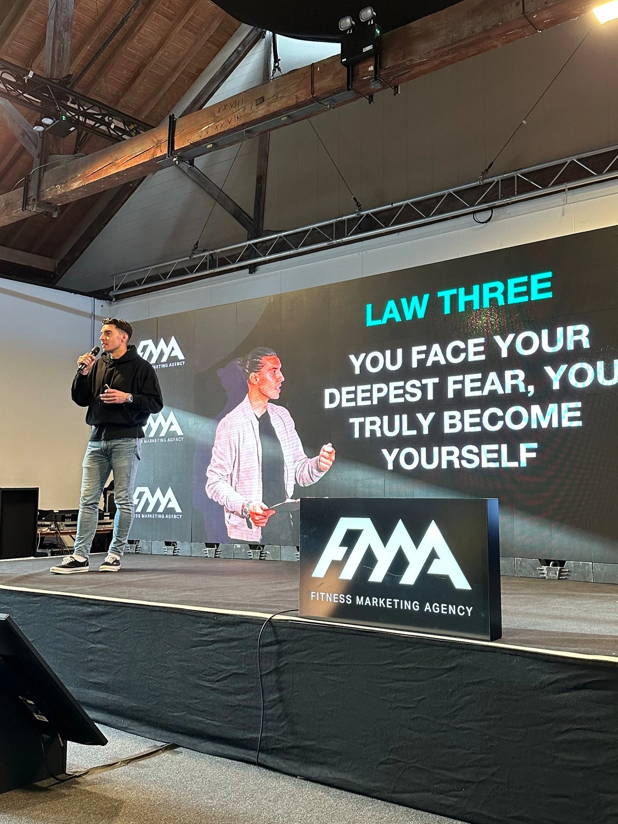 A person speaking on stage at a Fitness Marketing Agency event. A screen displays "Law Three: You face your deepest fear, you truly become yourself." The stage has a large sign with the agency's logo.